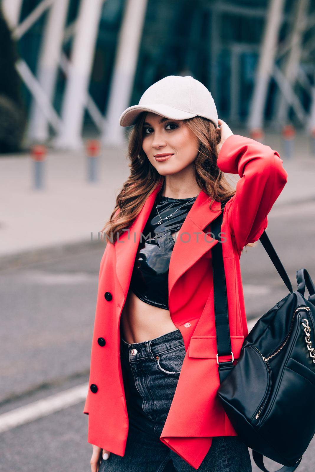 glamour woman in trendy outfit posing against the building urban background, fashion look. Outdoor fashion portrait of stylish young woman wearing black jeans, red jacket, top and a cap