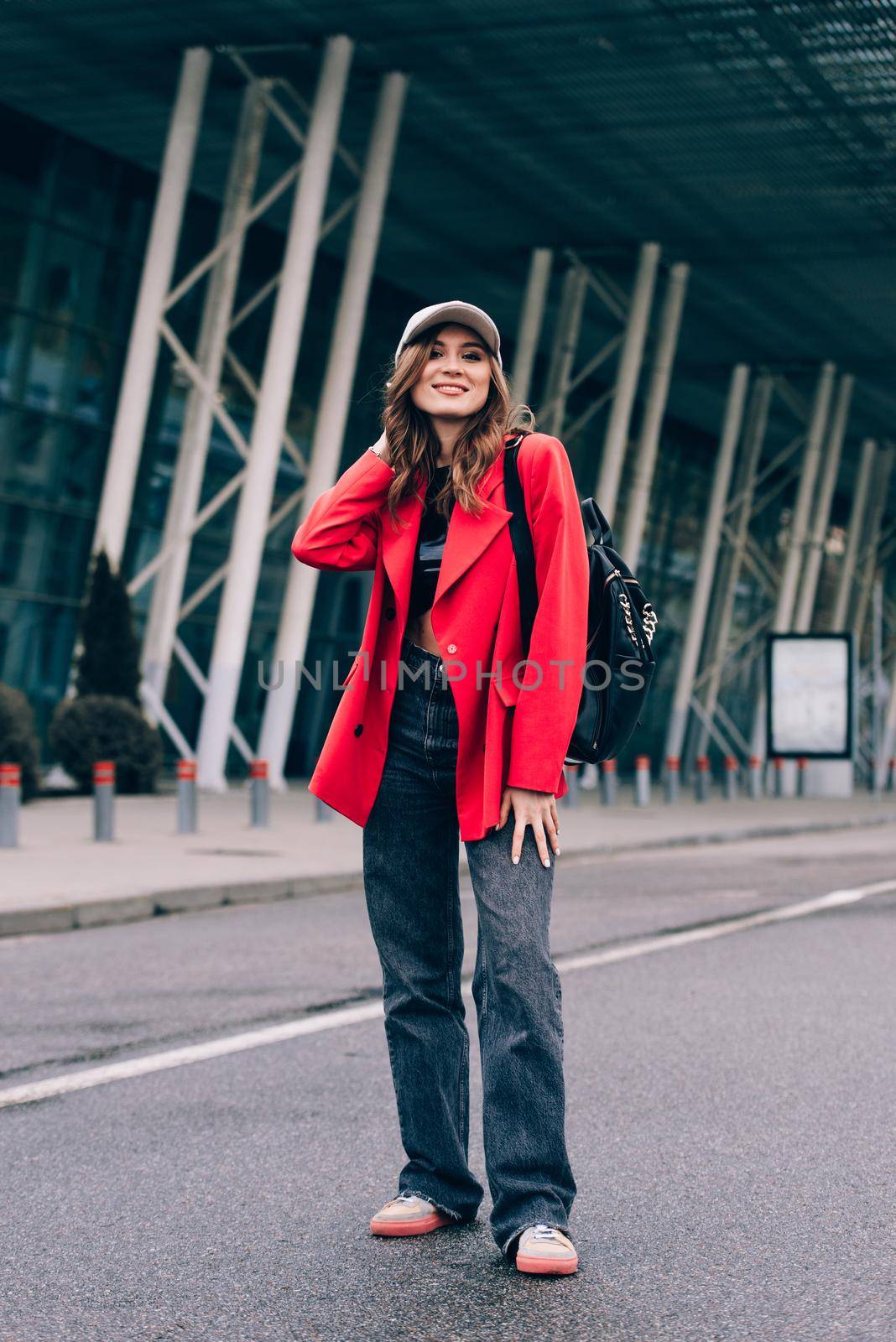 glamour woman in trendy outfit posing against the building urban background, fashion look. Outdoor fashion portrait of stylish young woman wearing black jeans, red jacket, top and a cap