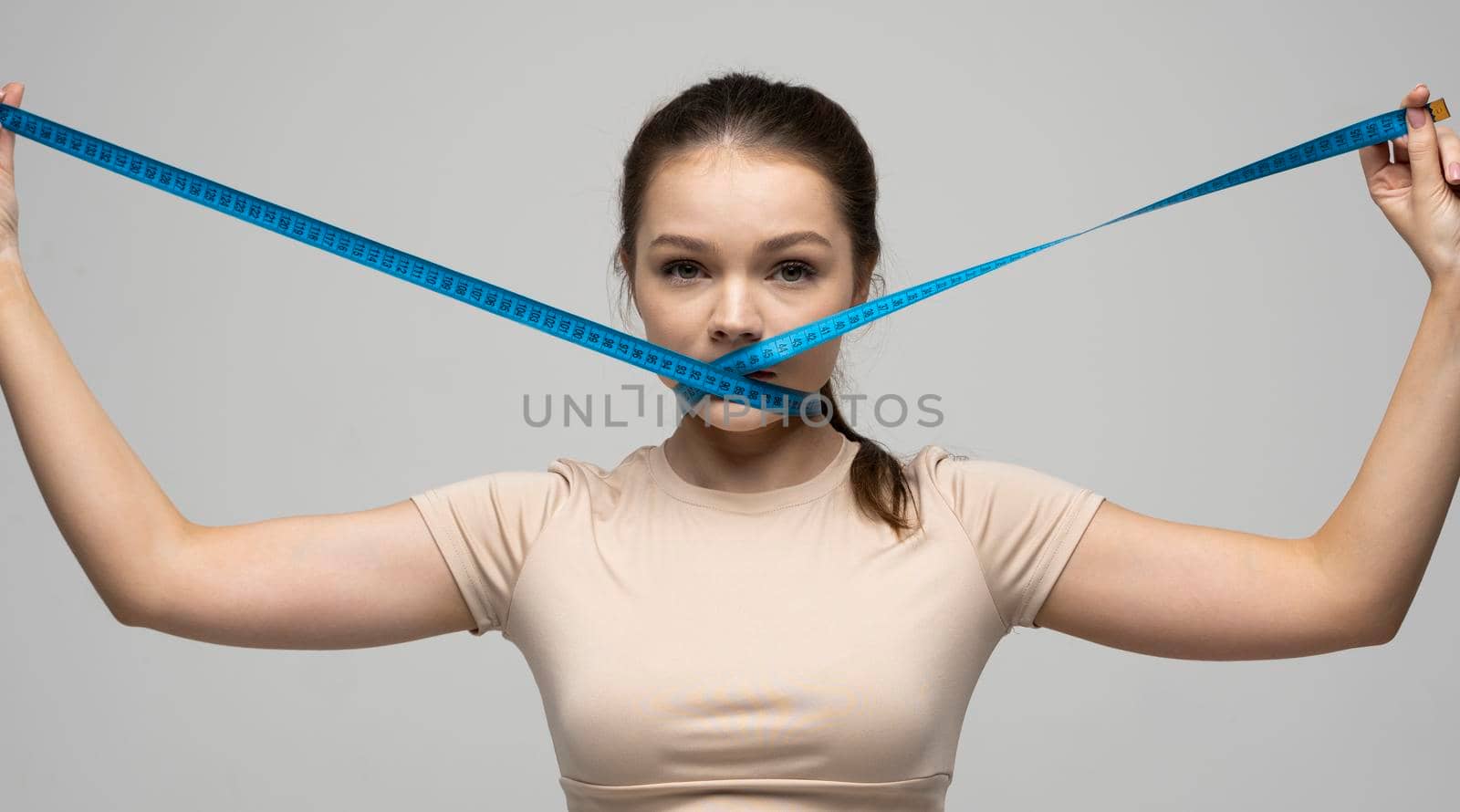 Young attractive beautiful brunette woman in a beige outfit holding measuring tape around her mouth on white background. Healthy lifestyle and diet concept