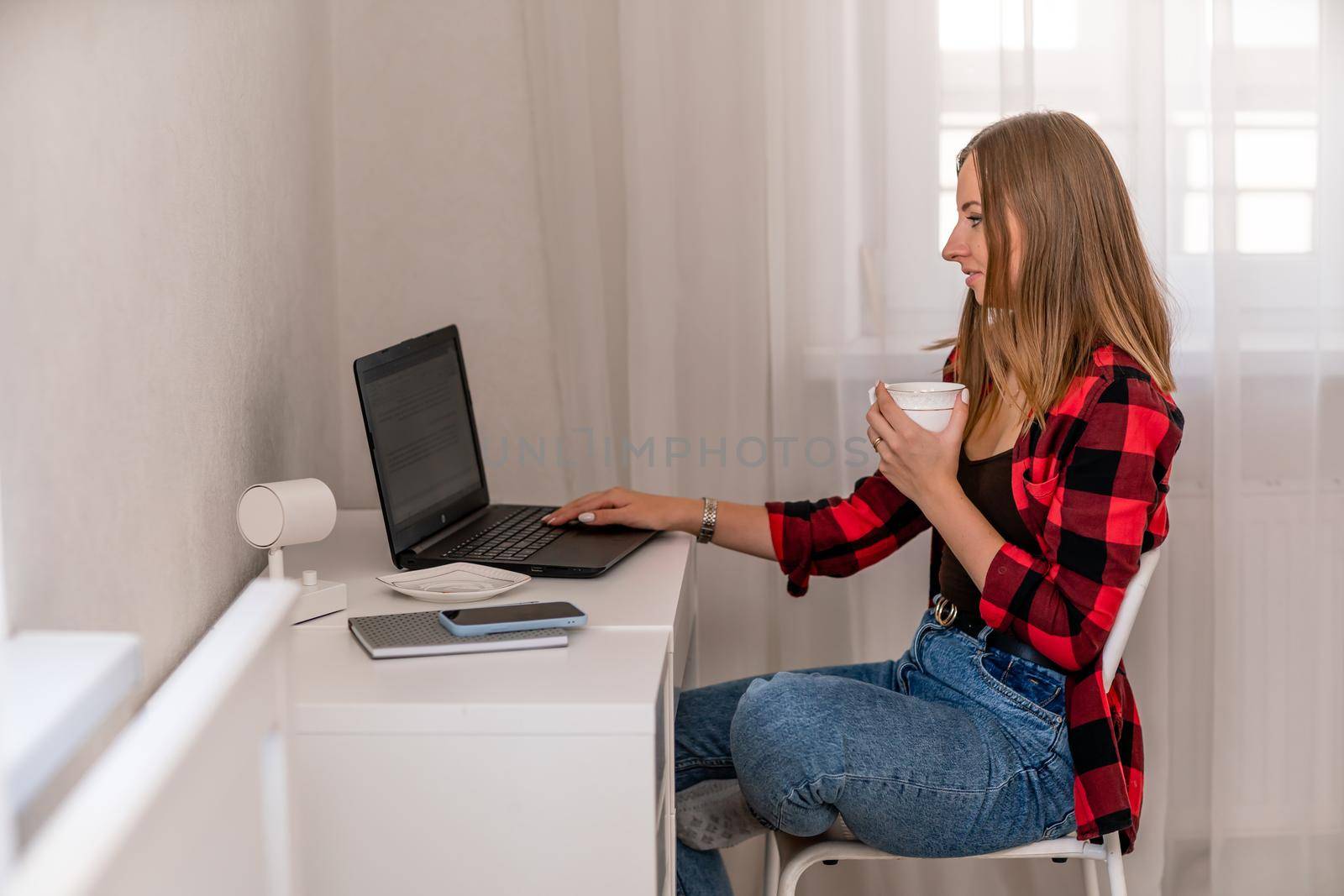 European professional woman sitting with laptop at home office desk, positive woman studying while working on PC. She is wearing a red plaid shirt and jeans