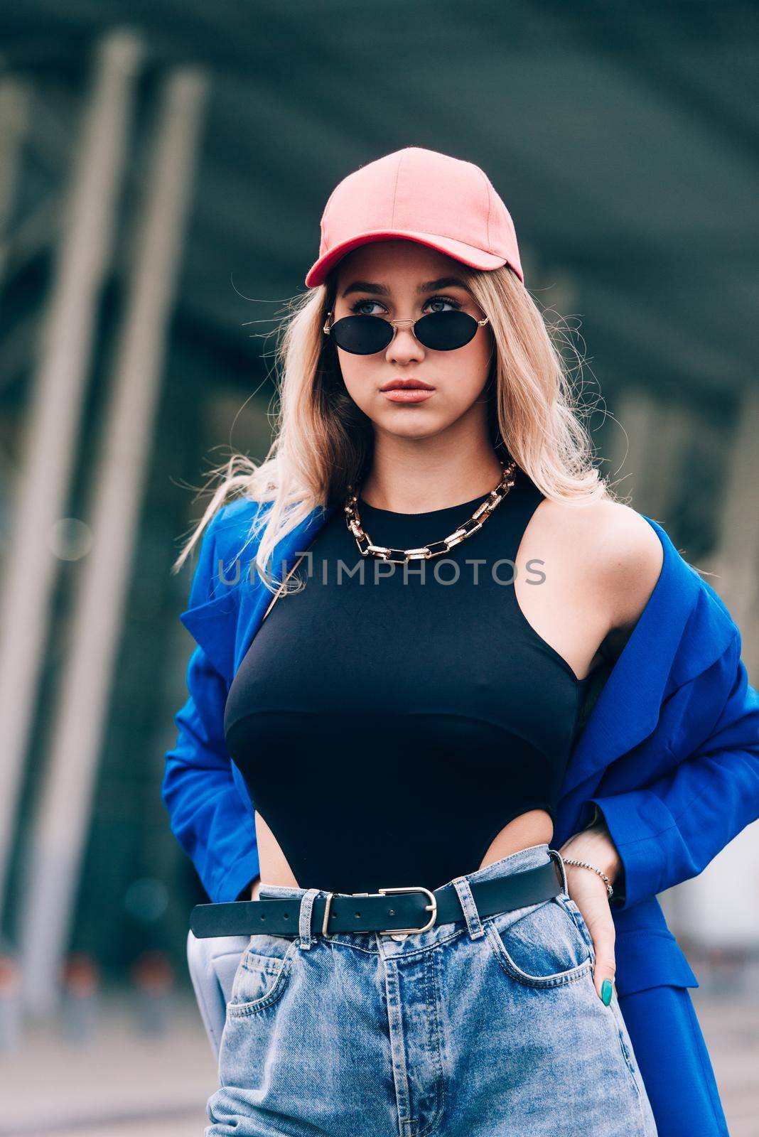 Young sexy blonde hipster woman posing on the street. Wearing blue stylish jacket, jeans and baseball hat and sunglasses. Lifestyle portrait