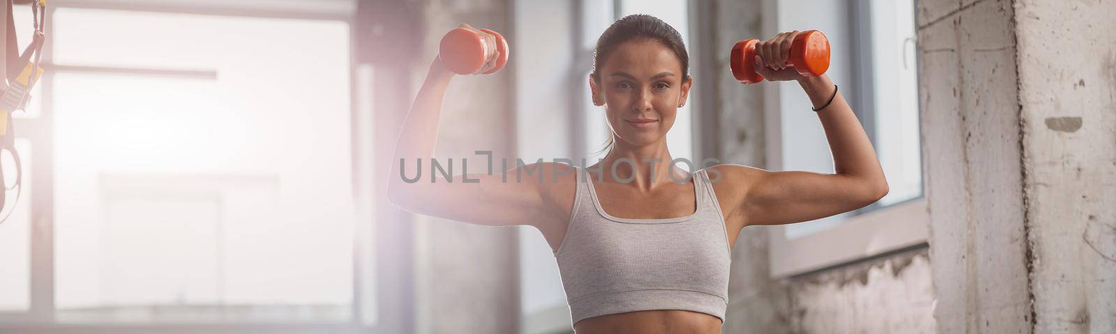 Young sporty woman holding dumbbells and performing exercise for arms, looking at camera in the fitness club