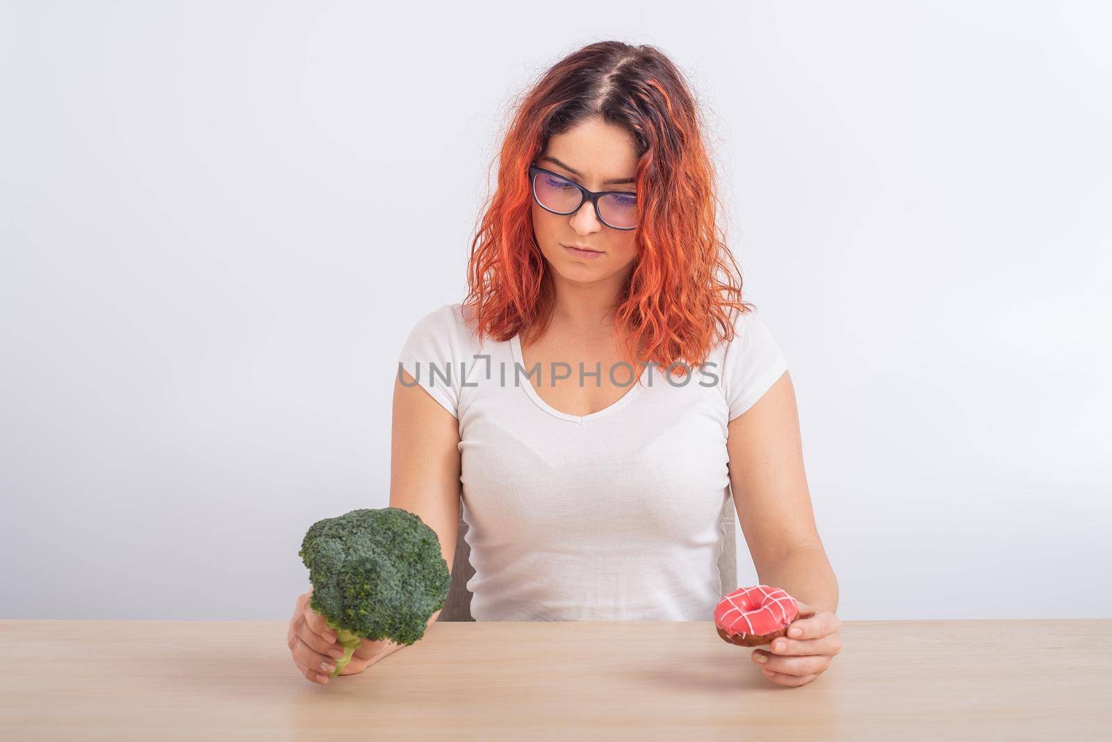 Caucasian woman chooses between vegetables and fast food. Redhead girl holding broccoli and donut on a white background. by mrwed54