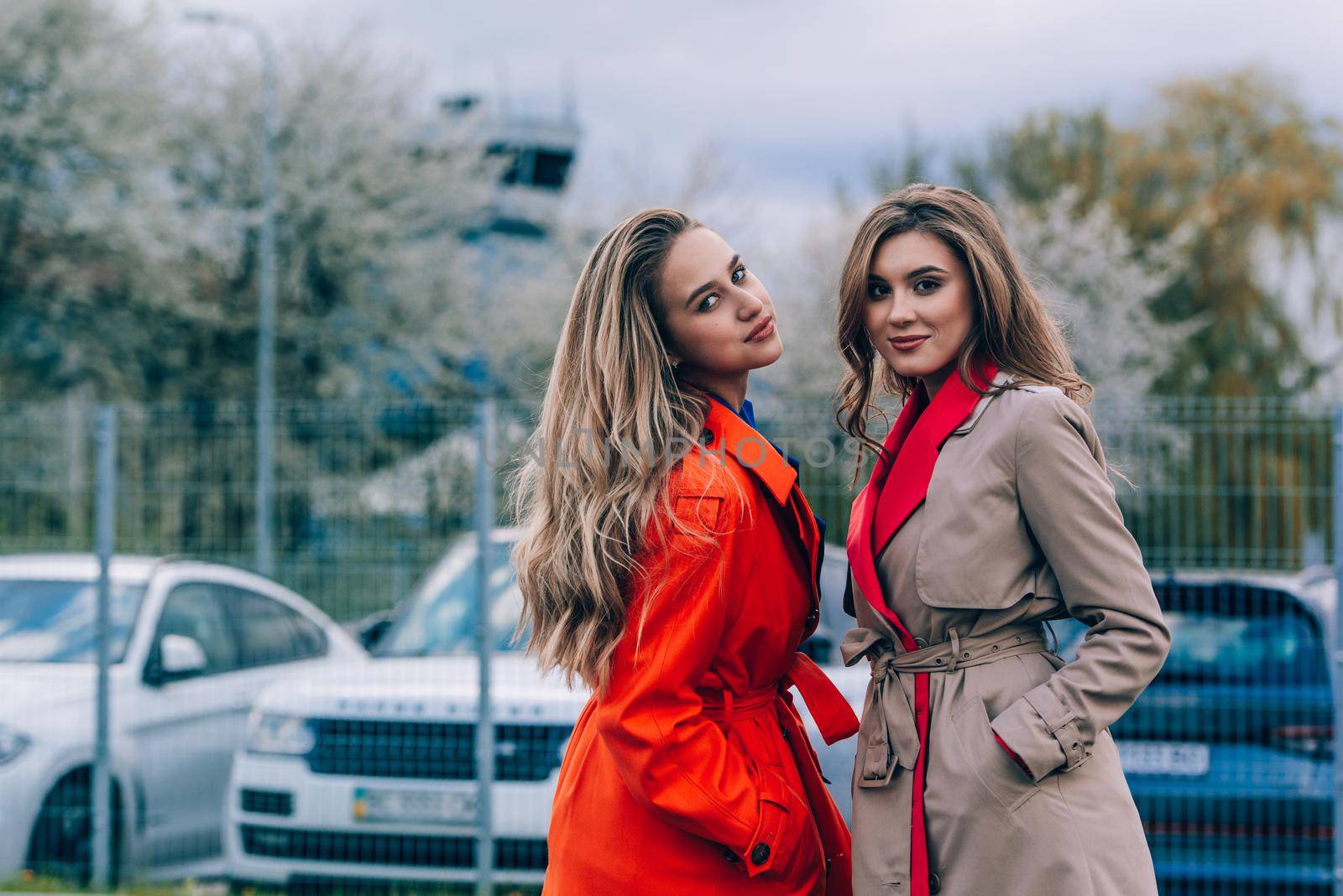 Fashionable happy smiling blonde woman wearing orange coat, blue jeanse and neckchain communicate with her girlfriend on the street. blonde and brunette happy and posing on the street