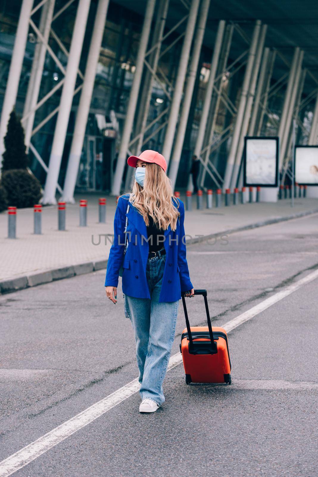 Portrait of a traveler woman in a mask walking with an orange suitcase near an airport. Young fashionable woman in a blue jeans and jacket by Ashtray25
