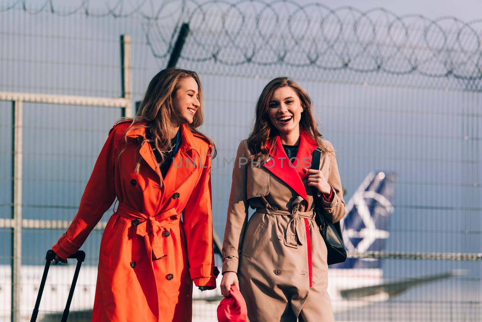 Two happy girls walking near airport, with luggage. Air travel, summer holiday. women dressed in trendy trenches orange and beige. Airport on a background