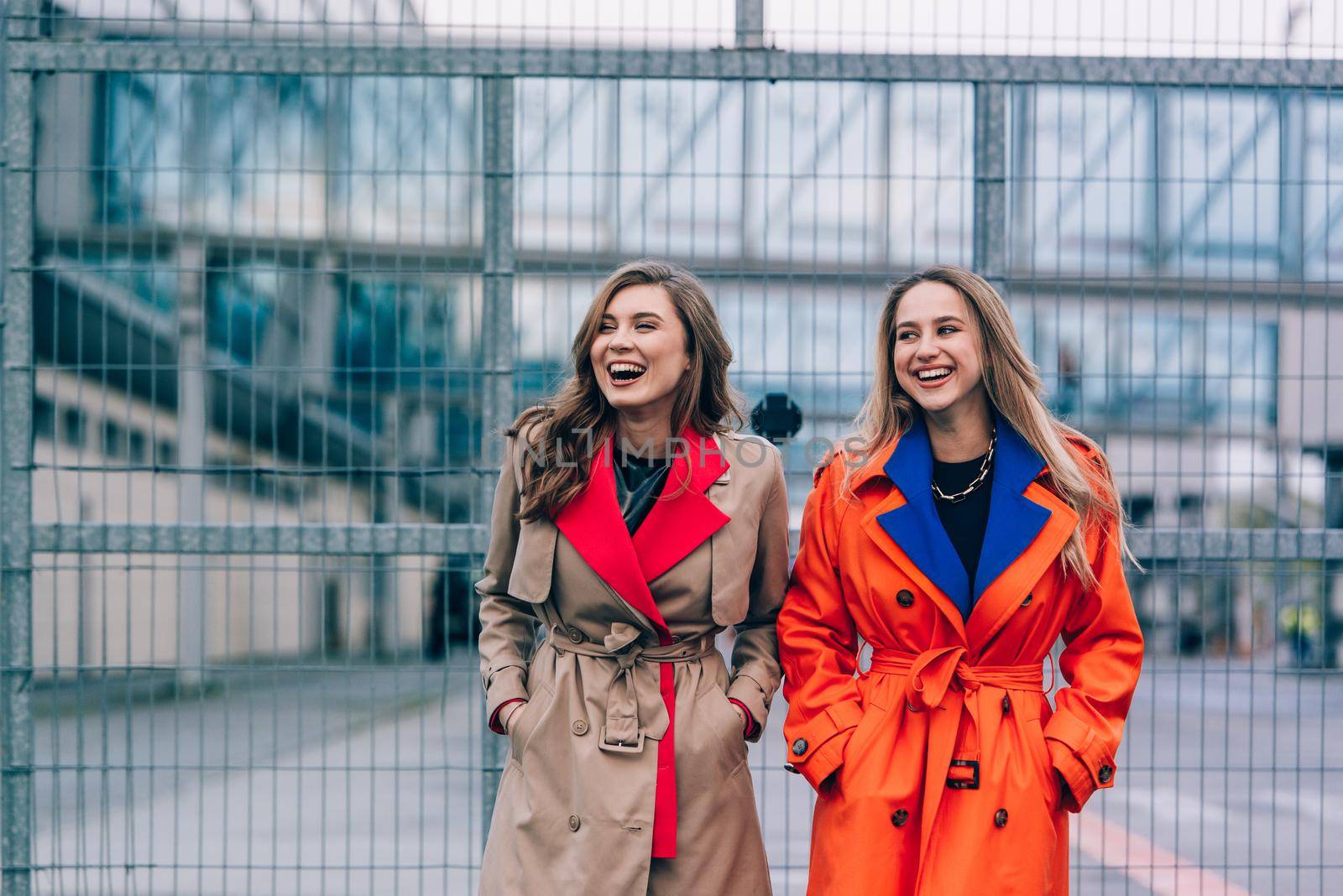 Fashionable happy smiling blonde woman wearing orange coat, blue jeanse and neckchain communicate with her girlfriend on the street. blonde and brunette happy and posing on the street