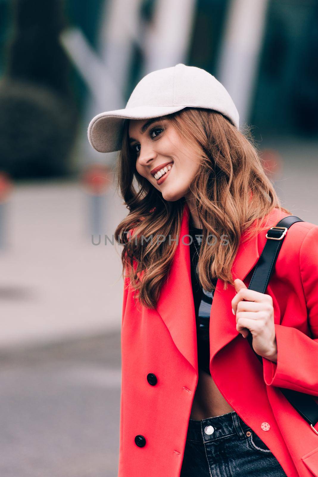 glamour woman in trendy outfit posing against the building urban background, fashion look. Outdoor fashion portrait of stylish young woman wearing black jeans, red jacket, top and a cap