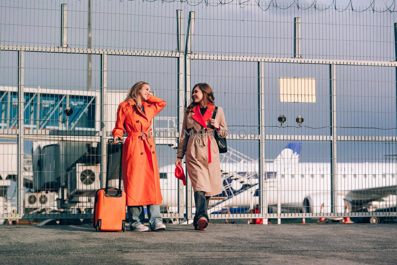 Two happy girls walking near airport, with luggage. Air travel, summer holiday. women dressed in trendy trenches orange and beige. Airport on a background