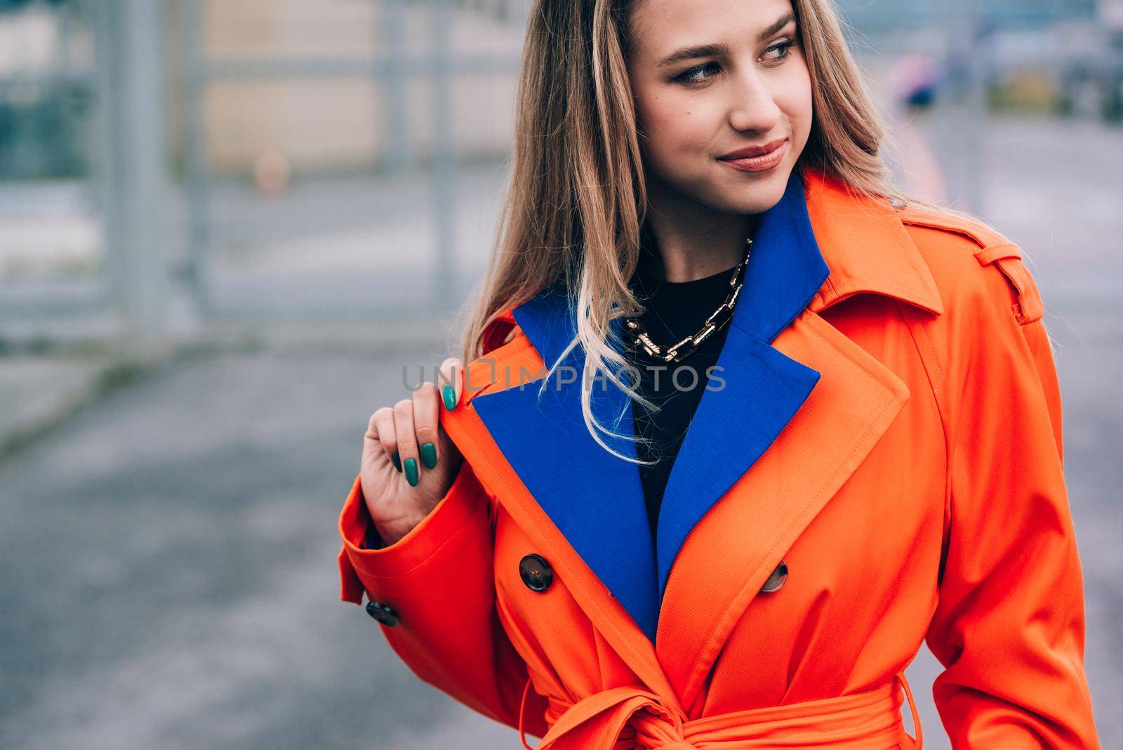 Fashionable blonde woman wearing orange coat, blue jeanse , white sneackers and neckchain posing in street of European city.
