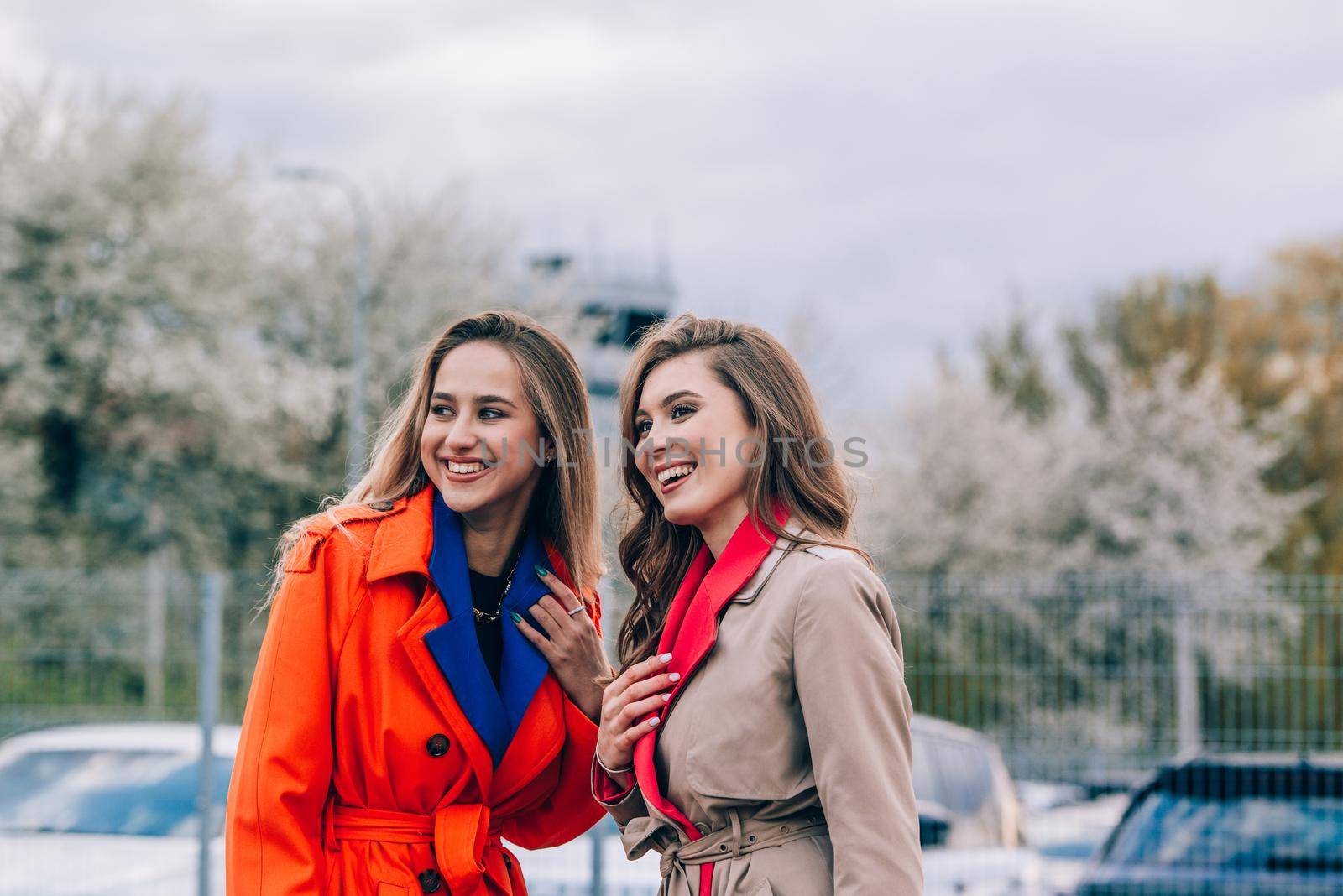 Fashionable happy smiling blonde woman wearing orange coat, blue jeanse and neckchain communicate with her girlfriend on the street. by Ashtray25
