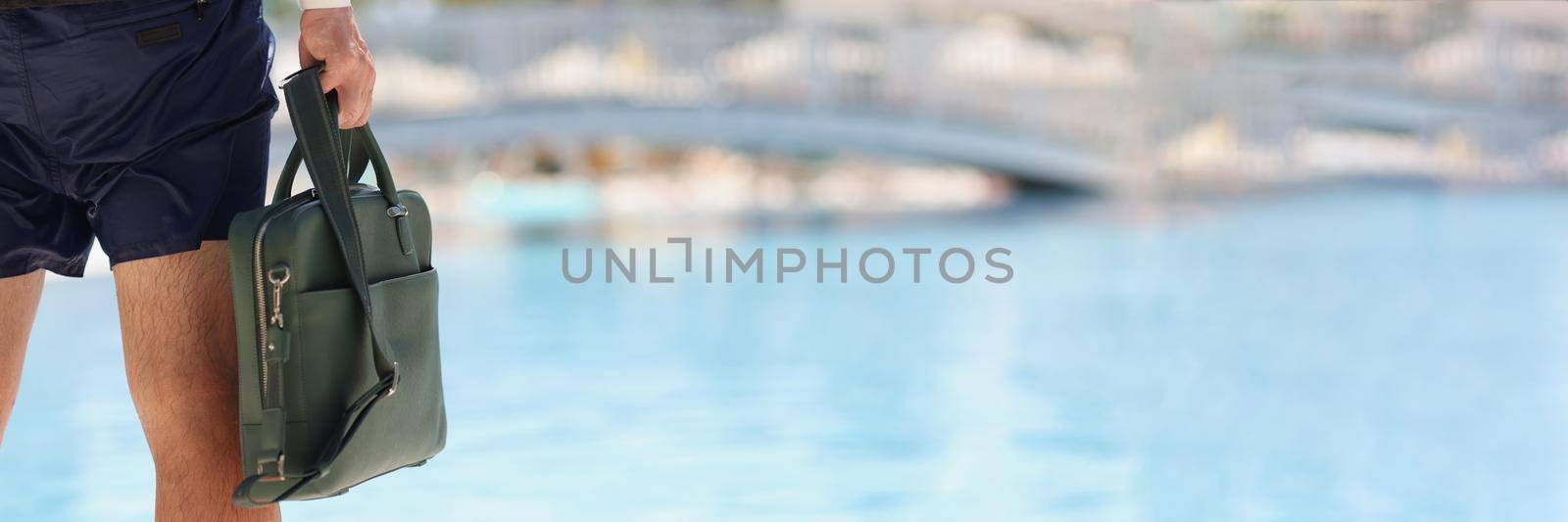 Close-up of man in wet pants holding bag, after swimming in pool. Business trip, time to go, end of vacation, summertime. Copy space in right. Blurred background