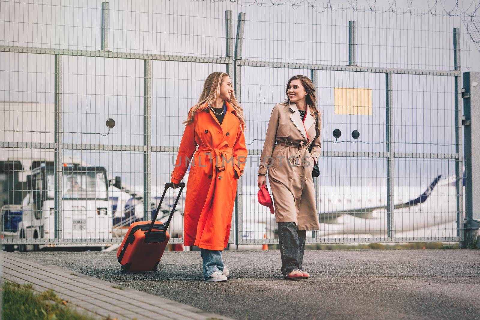 Two happy girls walking near airport, with luggage. Air travel, summer holiday. women dressed in trendy trenches orange and beige. Airport on a background