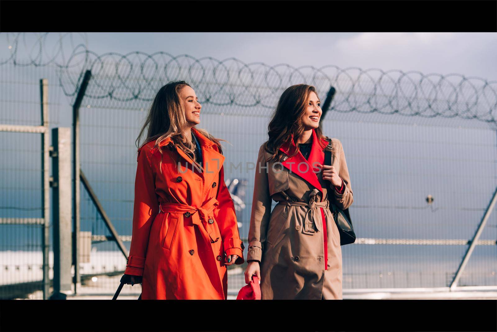 Two happy girls walking near airport, with luggage. Air travel, summer holiday. women dressed in trendy trenches orange and beige. Airport on a background