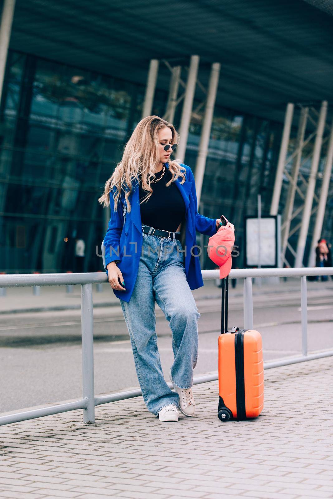 Portrait of a traveler woman standing with an orange suitcase near an airport. Young fashionable woman in a blue jeans and jacket, black shirt and white sneakers by Ashtray25