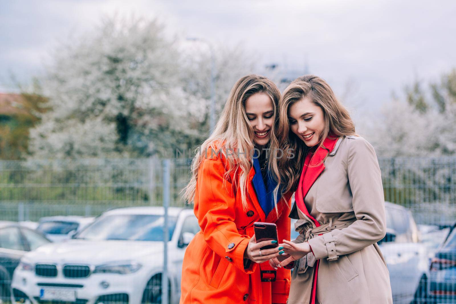 Fashionable happy smiling blonde woman wearing orange coat, blue jeanse and neckchain communicate with her girlfriend on the street. by Ashtray25