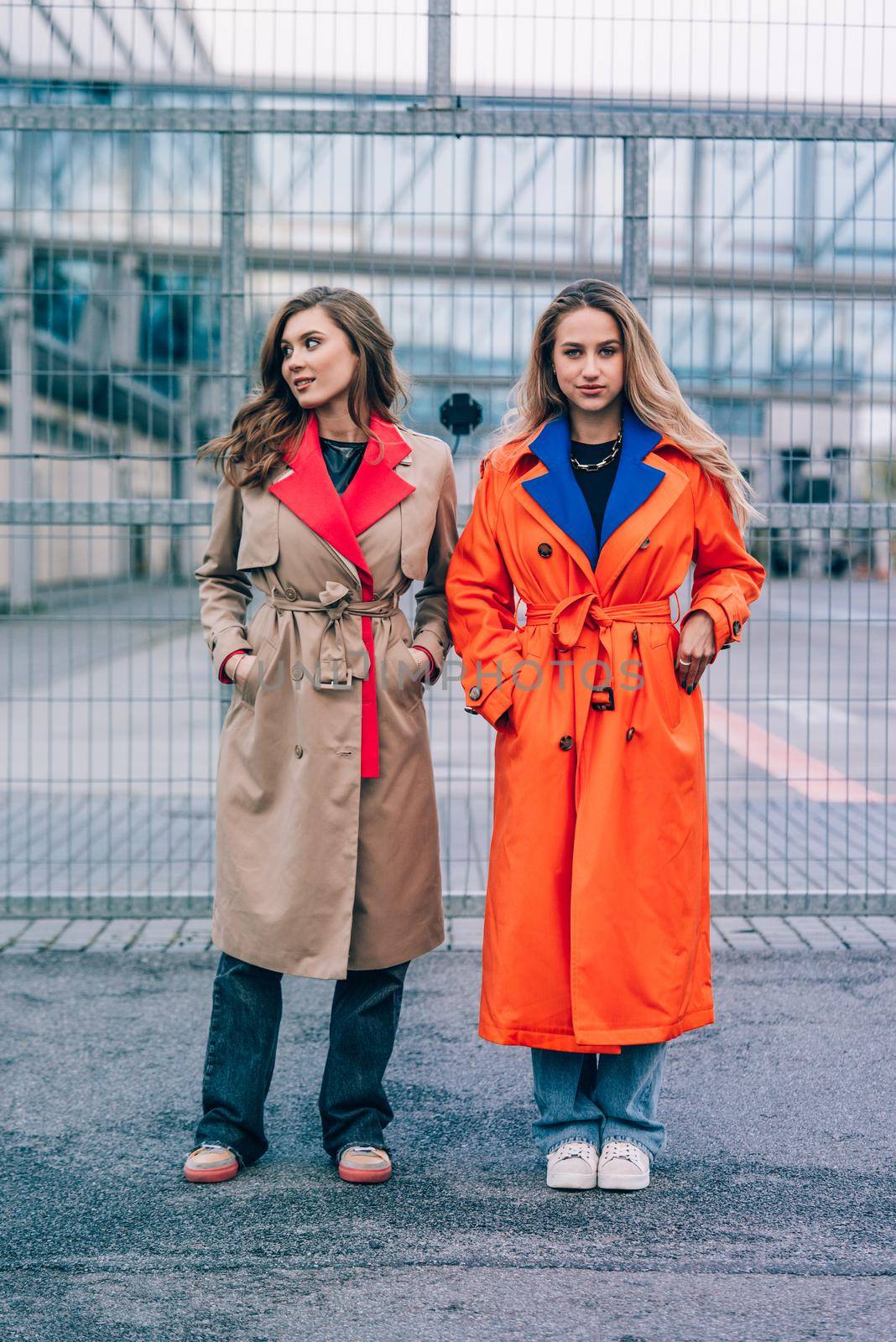 Fashionable happy smiling blonde woman wearing orange coat, blue jeanse and neckchain communicate with her girlfriend on the street. blonde and brunette happy and posing on the street