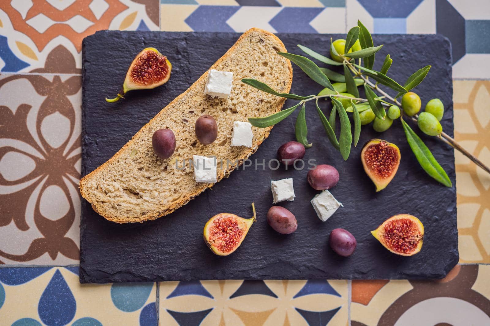 Green and black olives with loaf of fresh bread, feta cheese and young olives branch on olive wood chopping board over dark background by galitskaya