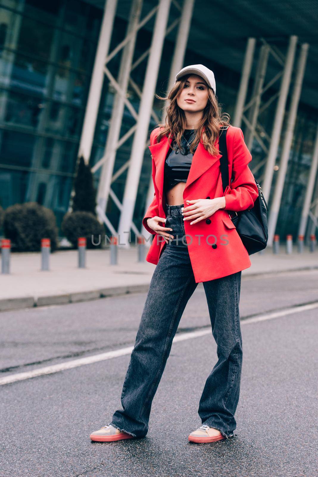 glamour woman in trendy outfit posing against the building urban background, fashion look. Outdoor fashion portrait of stylish young woman wearing black jeans, red jacket, top and a cap