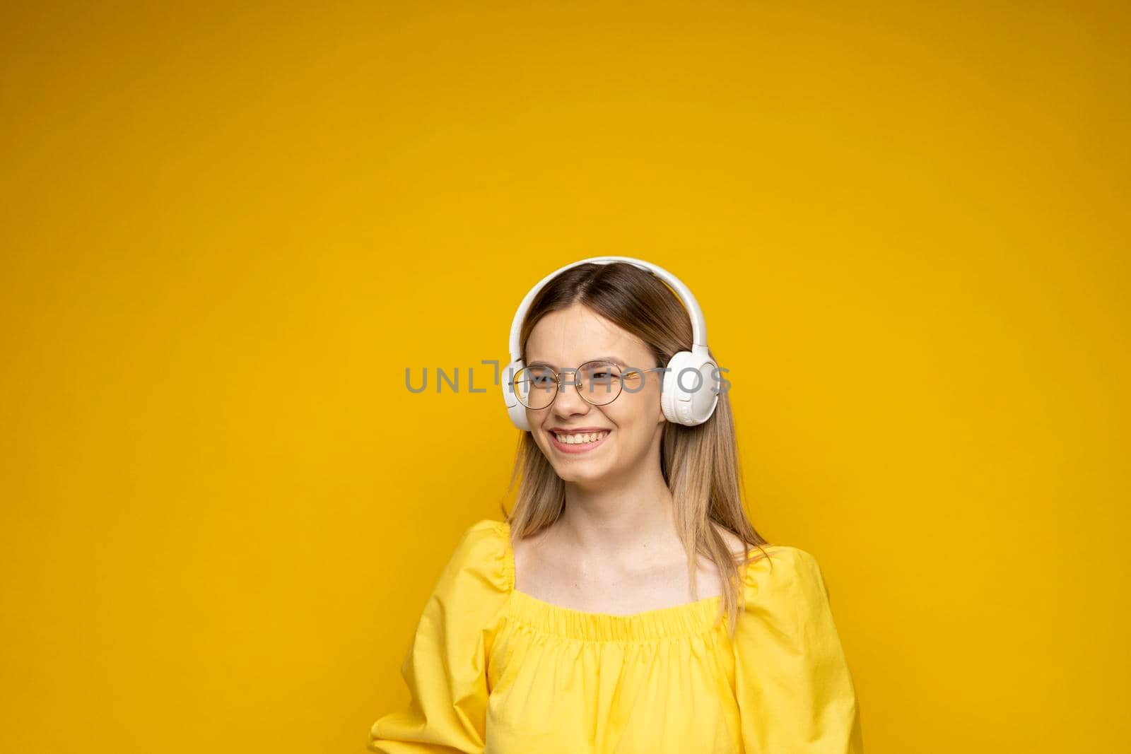 Beautiful young blonde woman in glasses with headphones listening to music and smiling and dancing, isolated on pink background