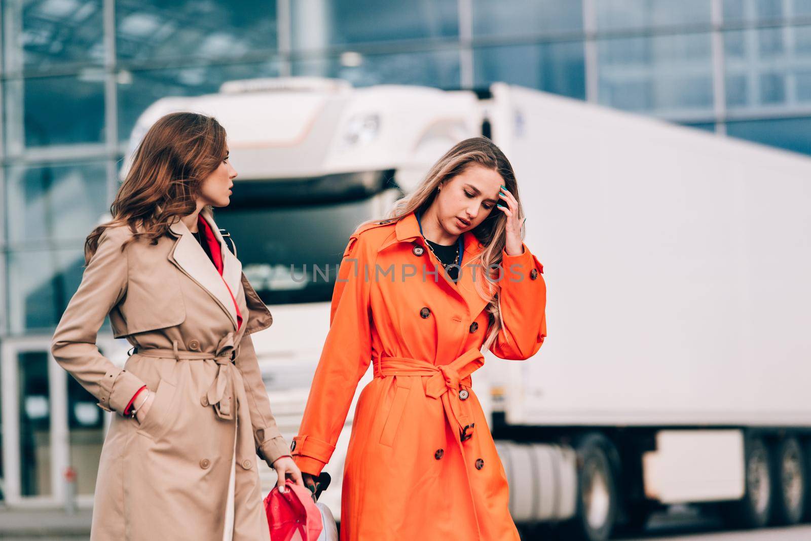 Two happy girls walking near airport, with luggage. Air travel, summer holiday. women dressed in trendy trenches orange and beige. Airport on a background
