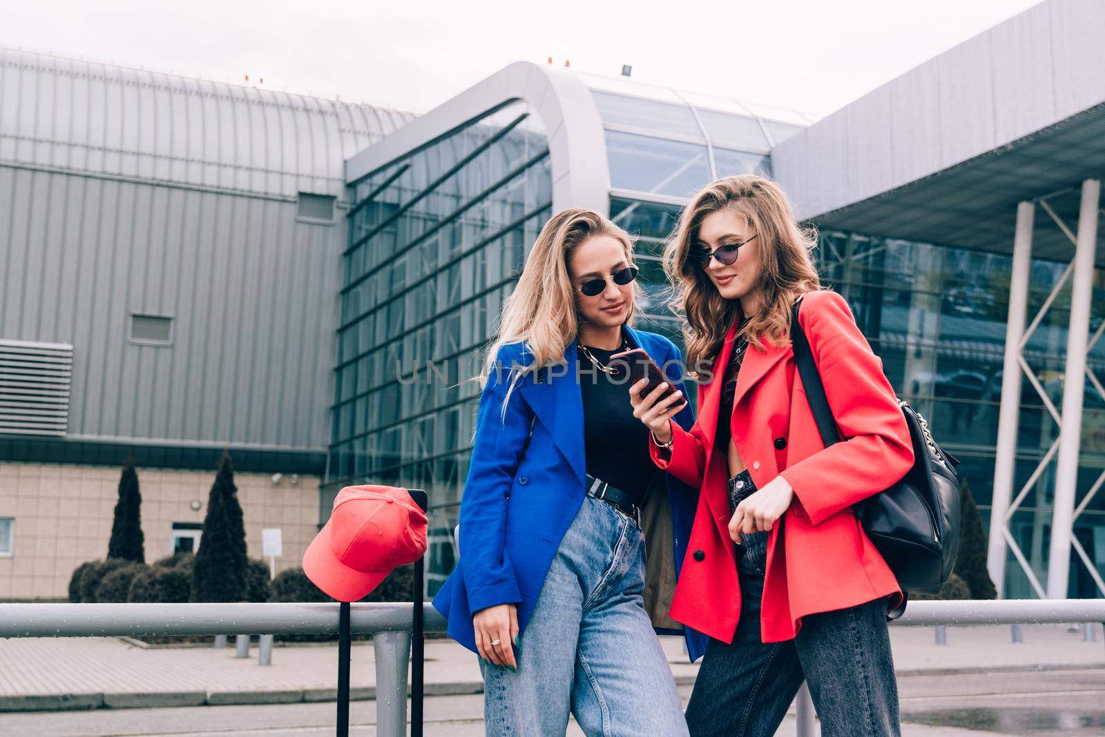 Two happy girls using smartphone checking flight or online check-in at airport together, with luggage. Air travel, summer holiday, or mobile phone application technology concept