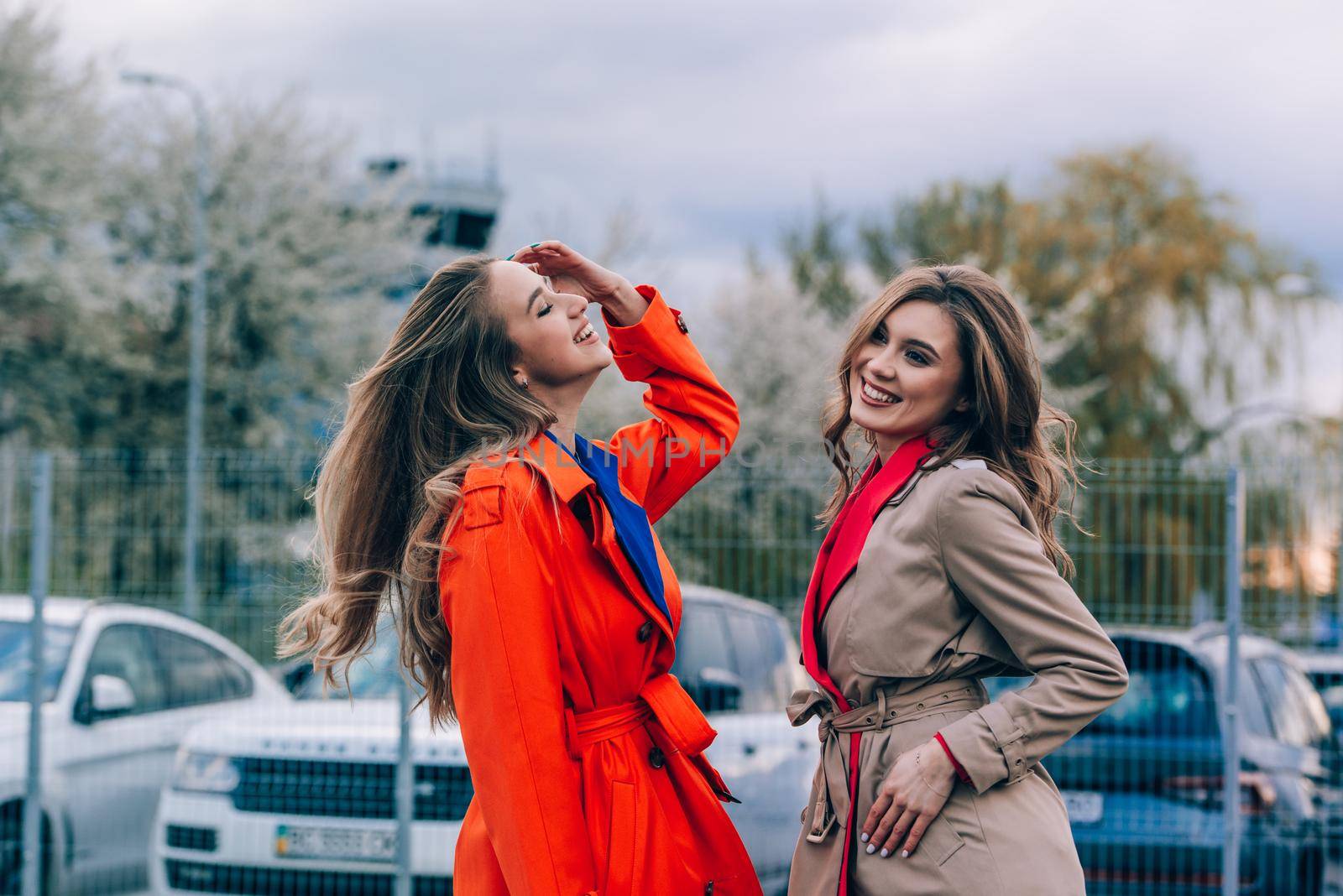 Fashionable happy smiling blonde woman wearing orange coat, blue jeanse and neckchain communicate with her girlfriend on the street. by Ashtray25