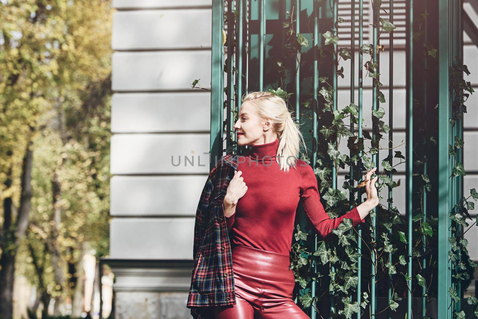 A beautiful, modern, fashionable blonde girl with a red lipstick posing outdoors . Dressed in a red leather leggings, turtleneck and checkered jacket.