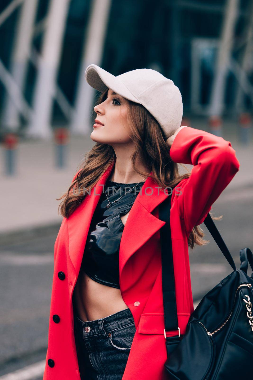 glamour woman in trendy outfit posing against the building urban background, fashion look. Outdoor fashion portrait of stylish young woman wearing black jeans, red jacket, top and a cap