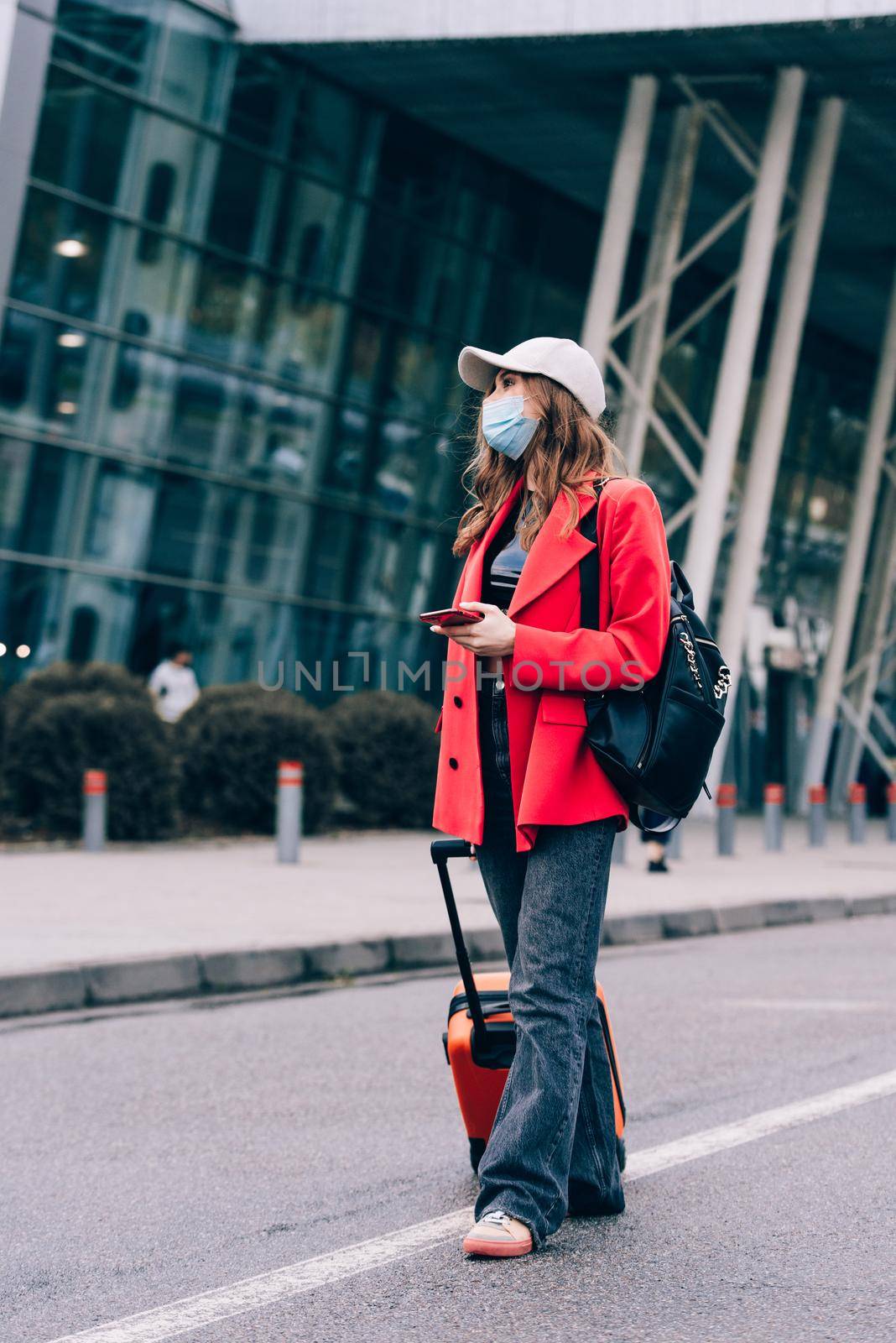 Portrait of a traveler woman in a mask walking with an orange suitcase near an airport. Young fashionable woman in a blue jeans and jacket by Ashtray25