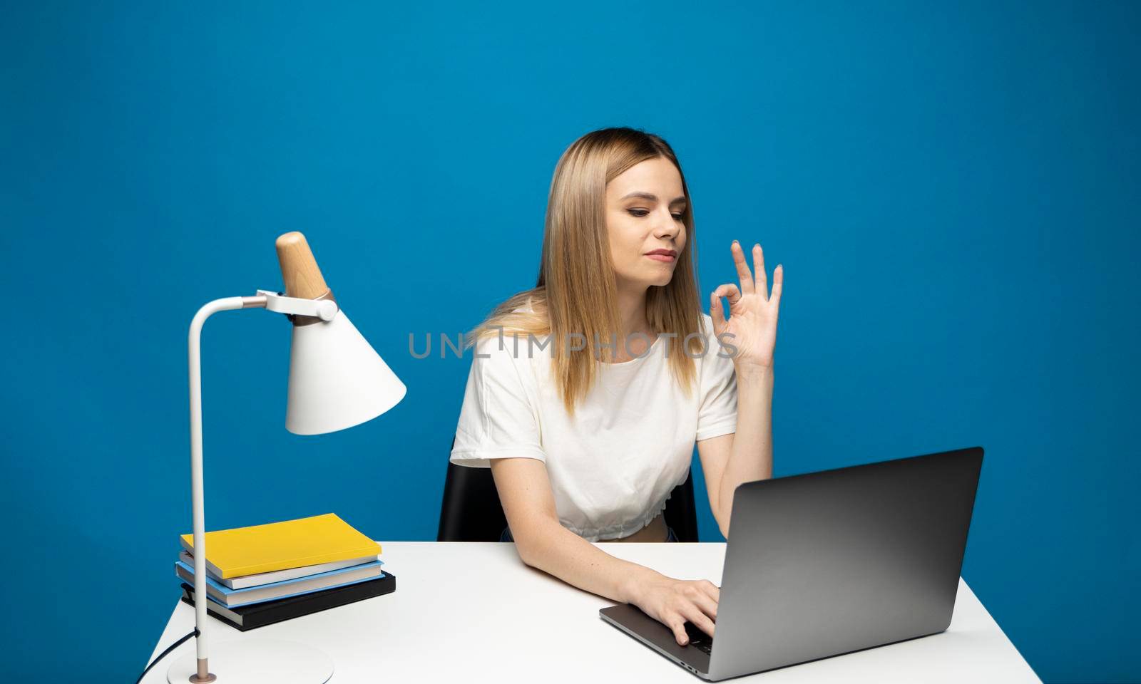 It's okay. Overjoyed woman in a white shirt and glasses showing ok sign, sitting with laptop over blue background. by vovsht