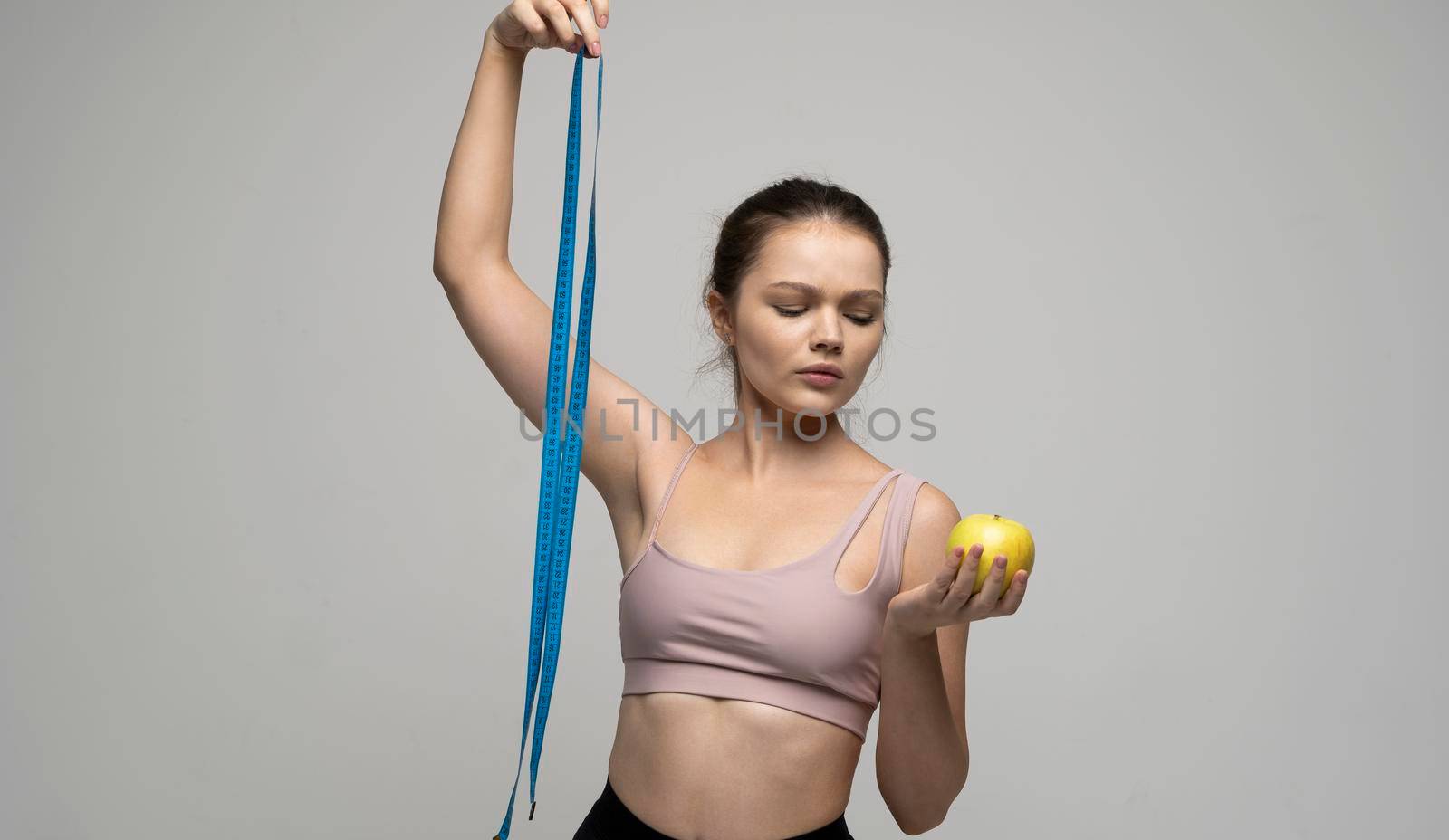 Confused attractive sexy brunette women in sport outfit standing with a measuring tape and holding a apple on a white background. Beauty and health concept. Healthy eating and diet. by vovsht