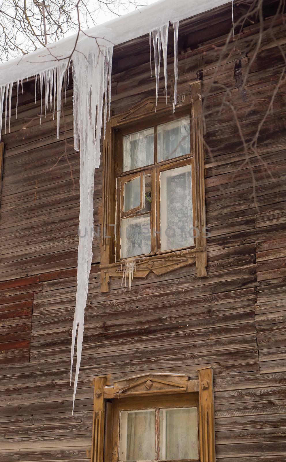 Icy transparent icicles hang on the edge of the roof. Against the background of the wooden wall of the old house. Large cascades, even beautiful rows. Cloudy winter day, soft light.