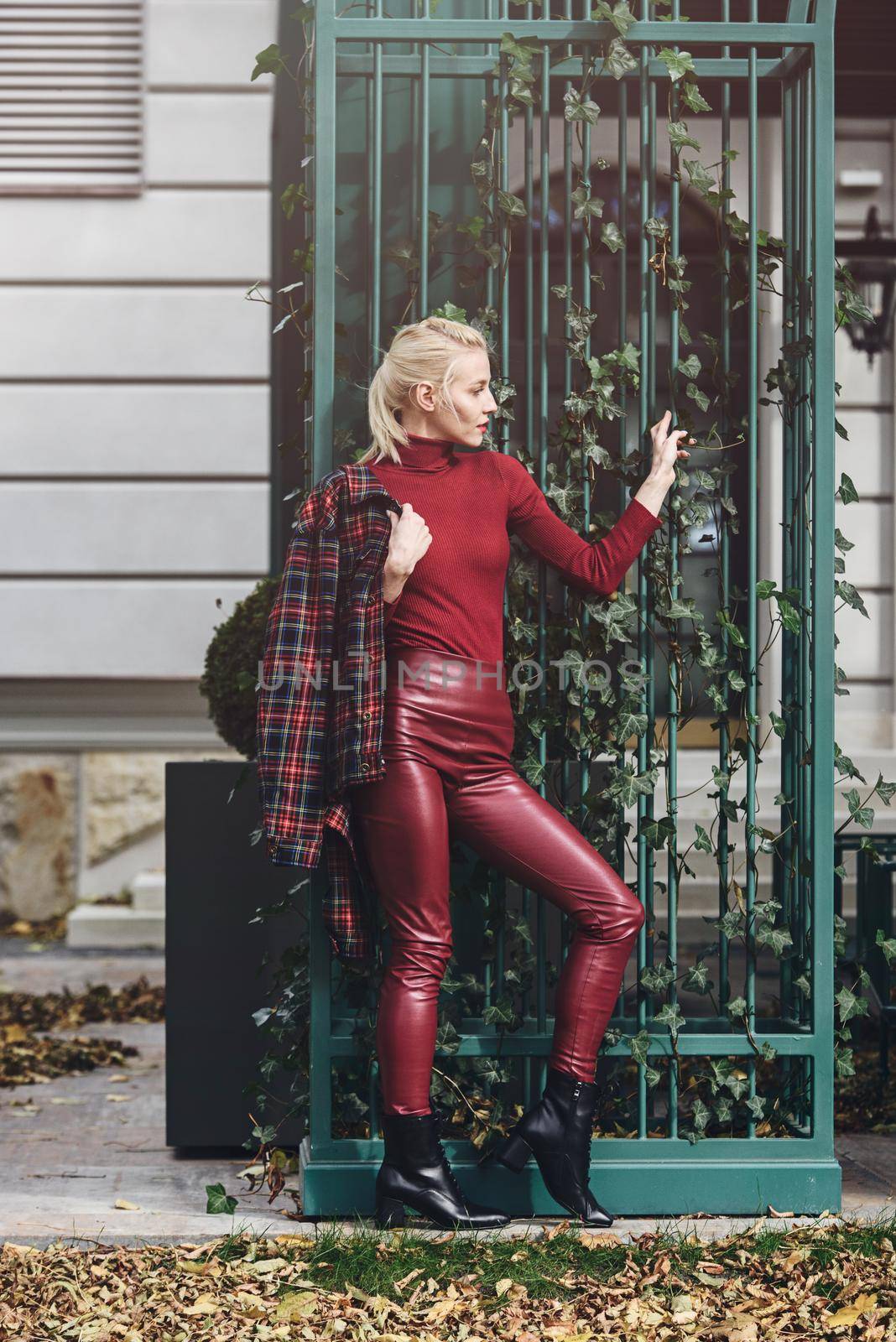 A beautiful, modern, fashionable blonde girl with a red lipstick posing outdoors . Dressed in a red leather leggings, turtleneck and checkered jacket.