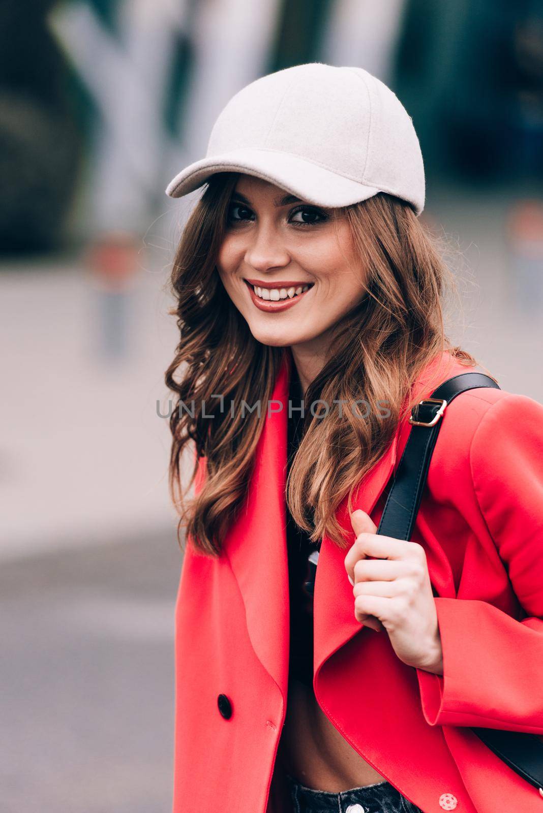 glamour woman in trendy outfit posing against the building urban background, fashion look. Outdoor fashion portrait of stylish young woman wearing black jeans, red jacket, top and a cap