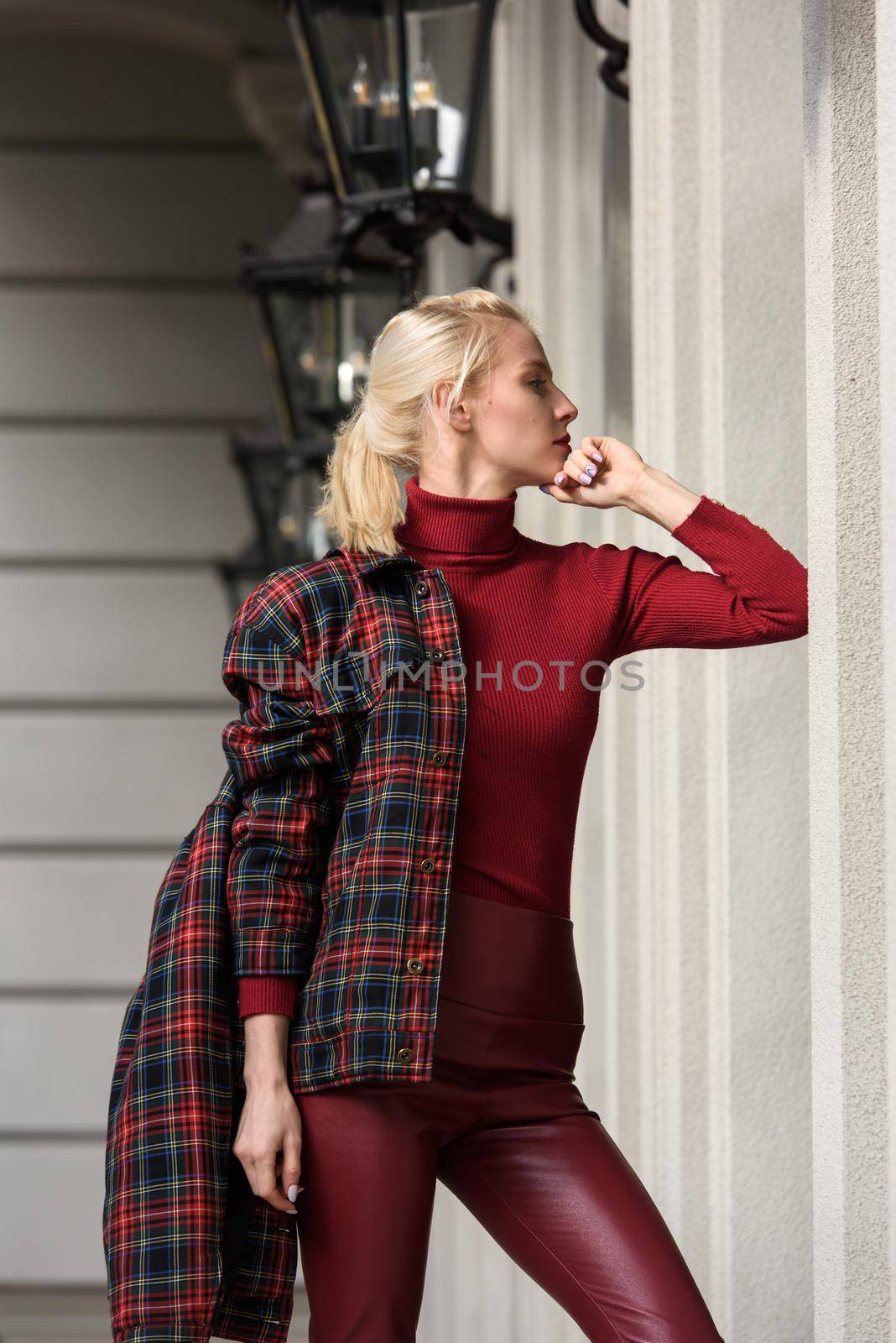 A beautiful, modern, fashionable blonde girl with a red lipstick posing outdoors . Dressed in a red leather leggings, turtleneck and checkered jacket.