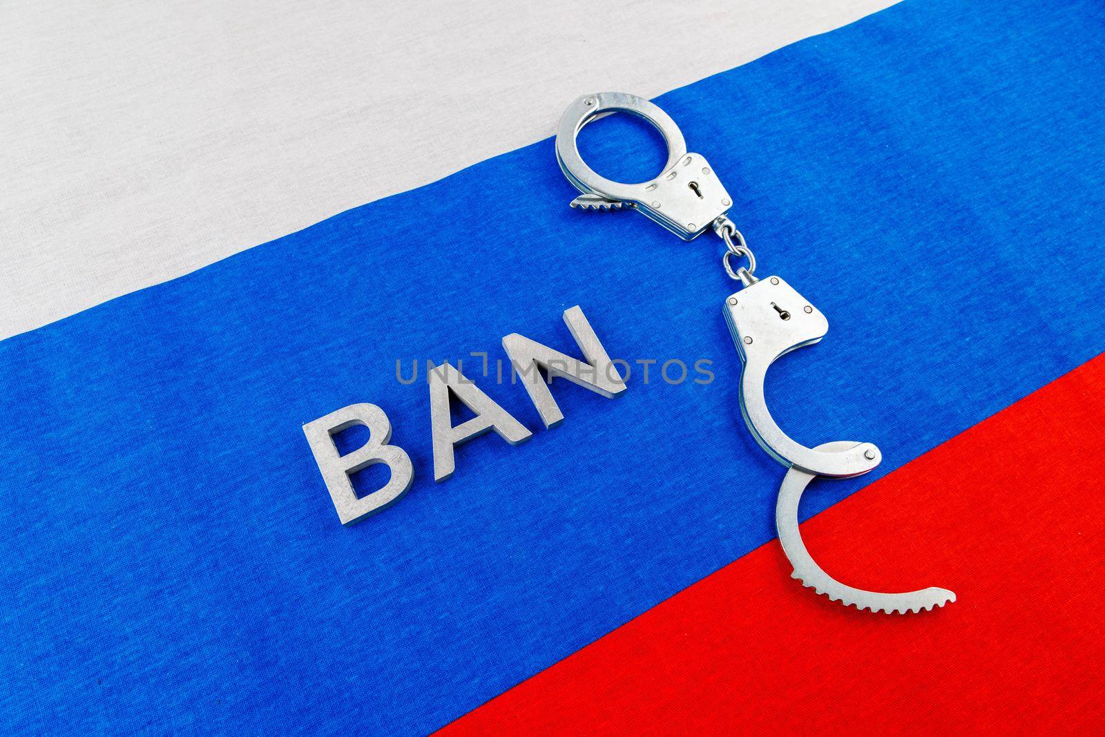 the word ban laid with silver metal letters near opened handcuffs over fabric tricolor flag of the Russian Federation