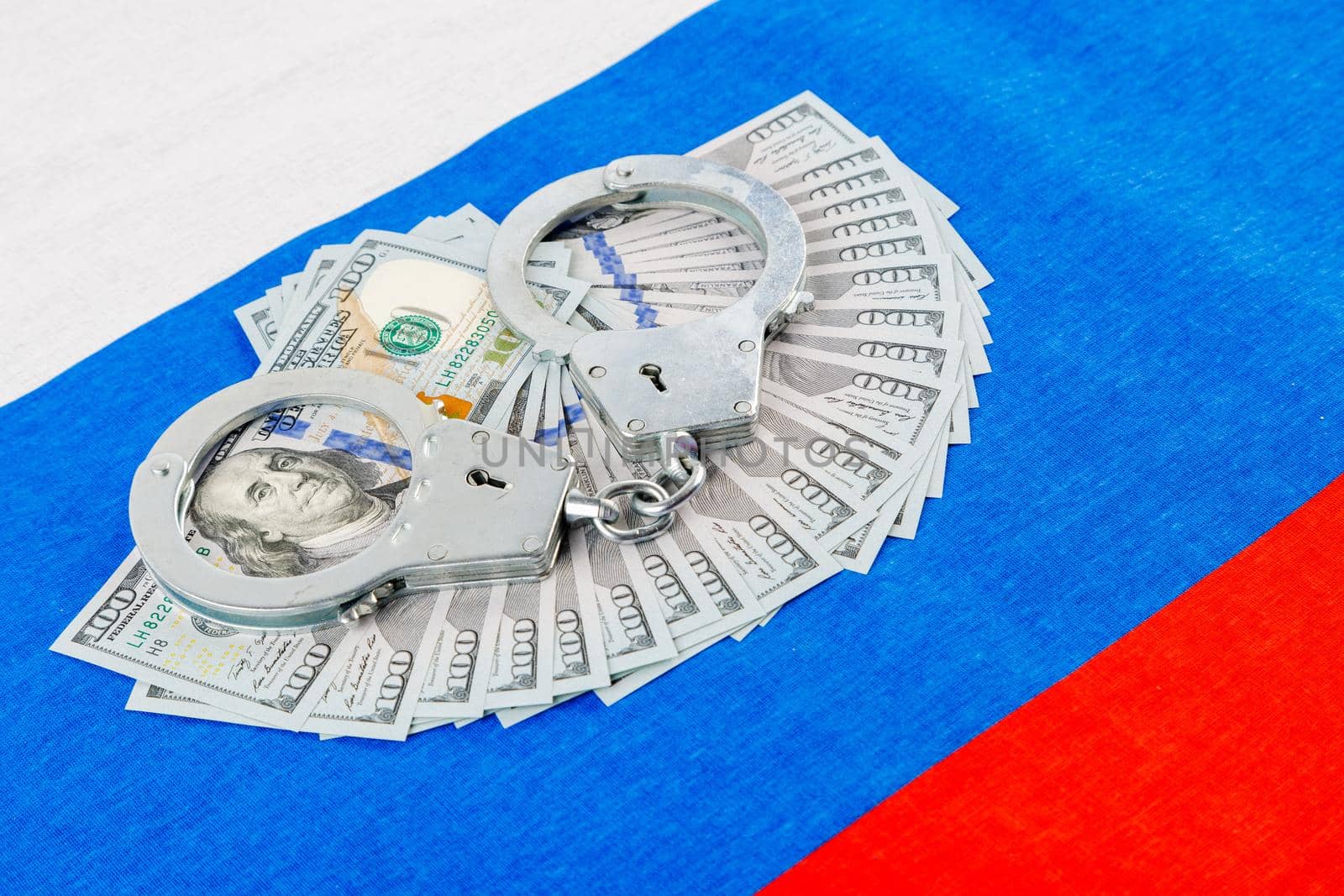 silver metal police handcuffs over paper dollar banknotes of United States of America over national flag of Russian Federation, concept of foreign currency prohibition