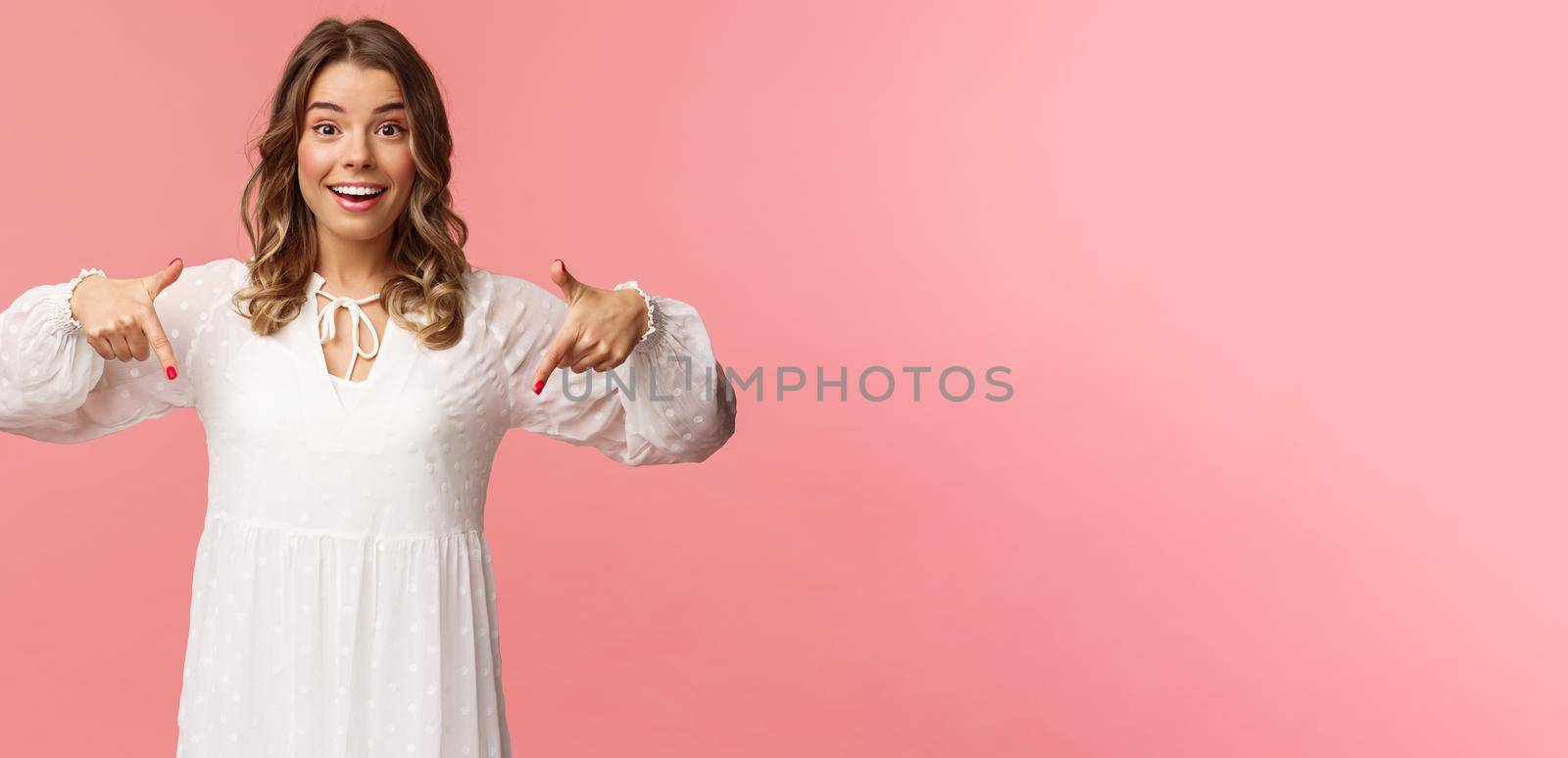 Portrait of enthusiastic upbeat young blond woman pointing fingers down to invite you check-out product, showing bottom advertisement, smiling camera cheerful, spring concept, pink background.