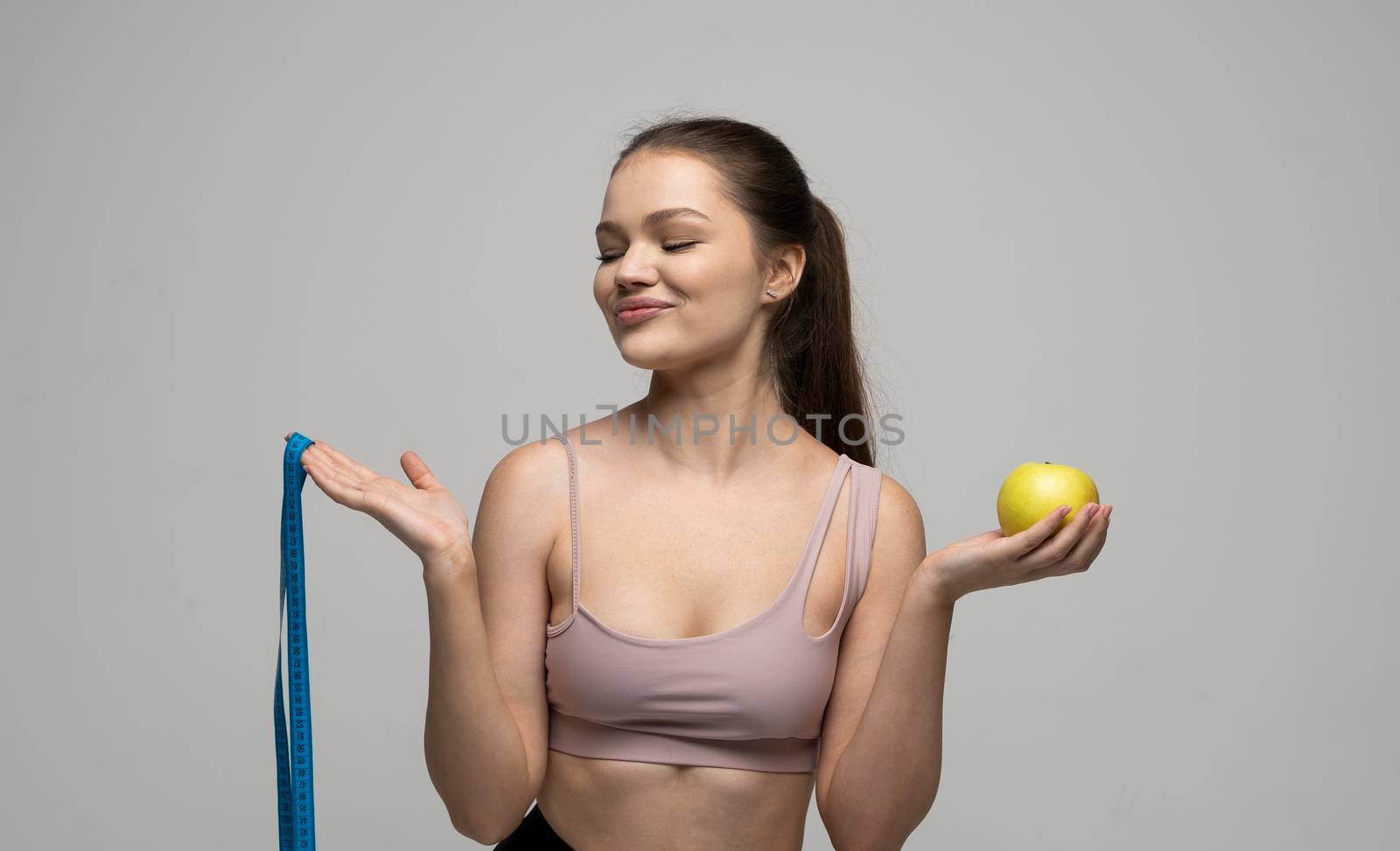 Brunette attractive woman in a sport wear is smile and looks in a camera while holds blue measuring tape and an apple. Health care and healthy nutrition. Perfect slim body