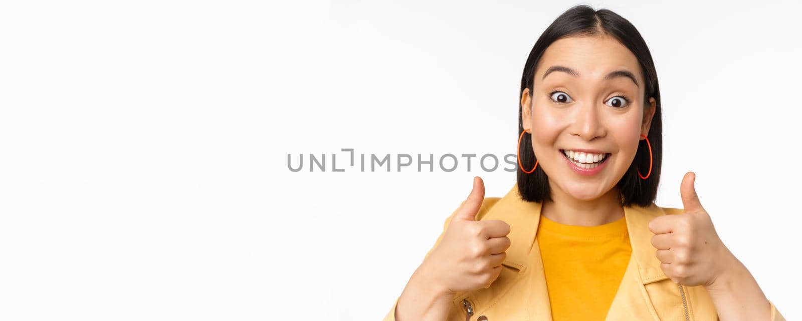 Smiling asian girl showing thumbs up, looking pleased, approve smth, standing over white background. Copy space