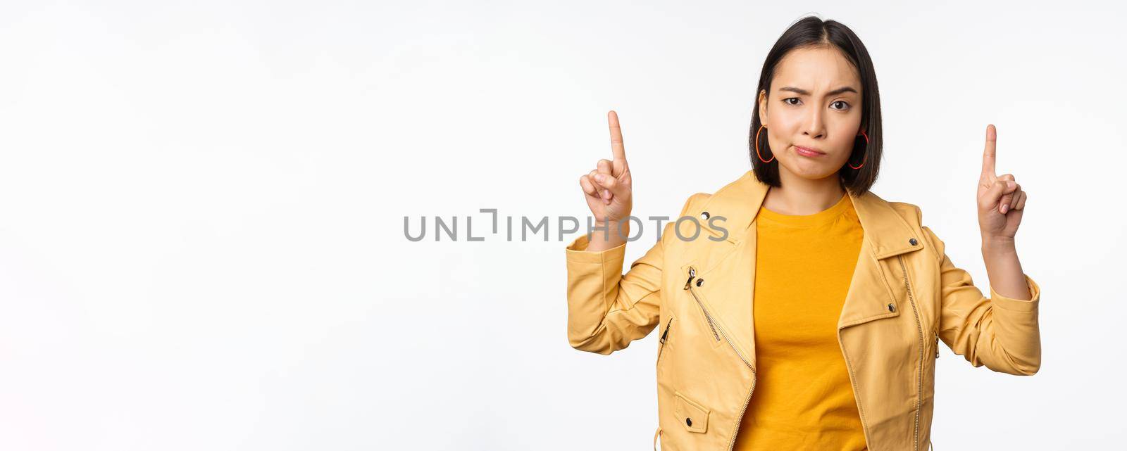 Skeptical asian woman pointing fingers up, frowning and looking with disapproval, complaining at smth, standing doubtful against white background.