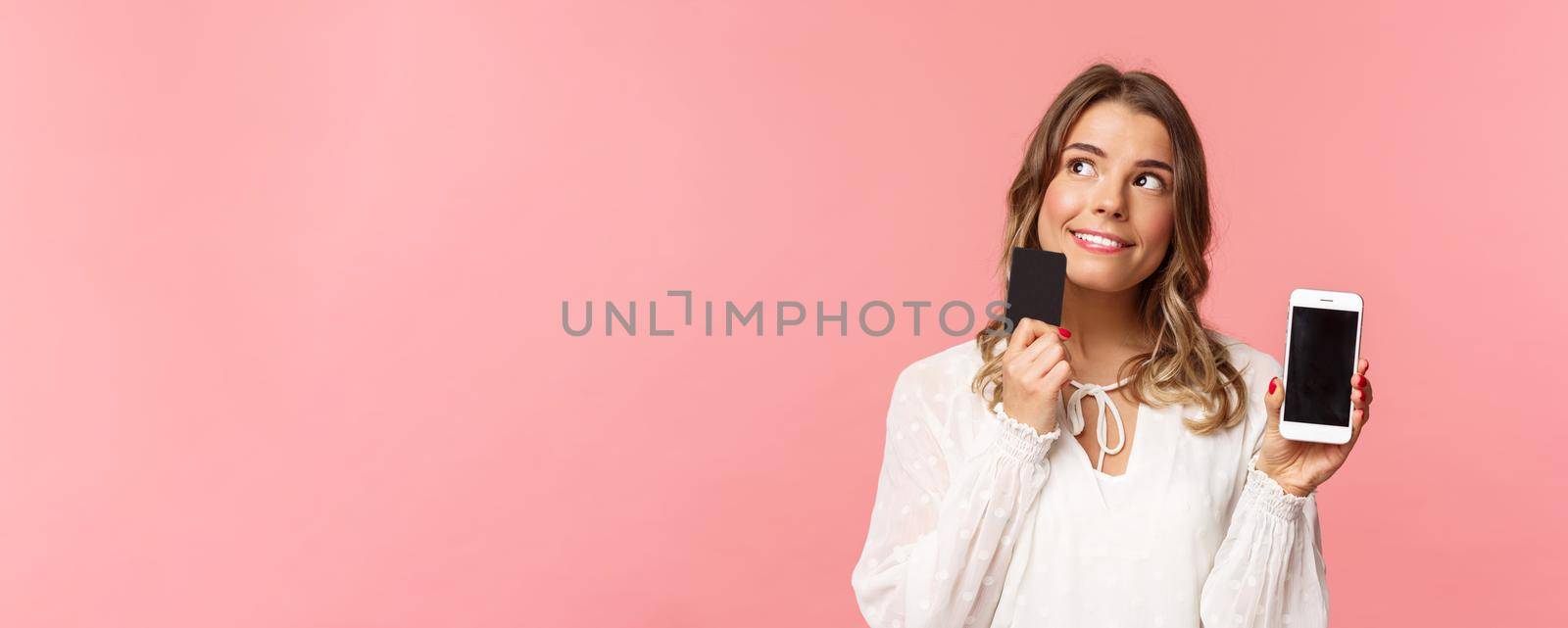 Finance, shopping and technology concept. Close-up portrait of thoughtful, dreamy attractive blond girl in white dress, thinking what buy, order something online shop, hold smartphone and credit card.