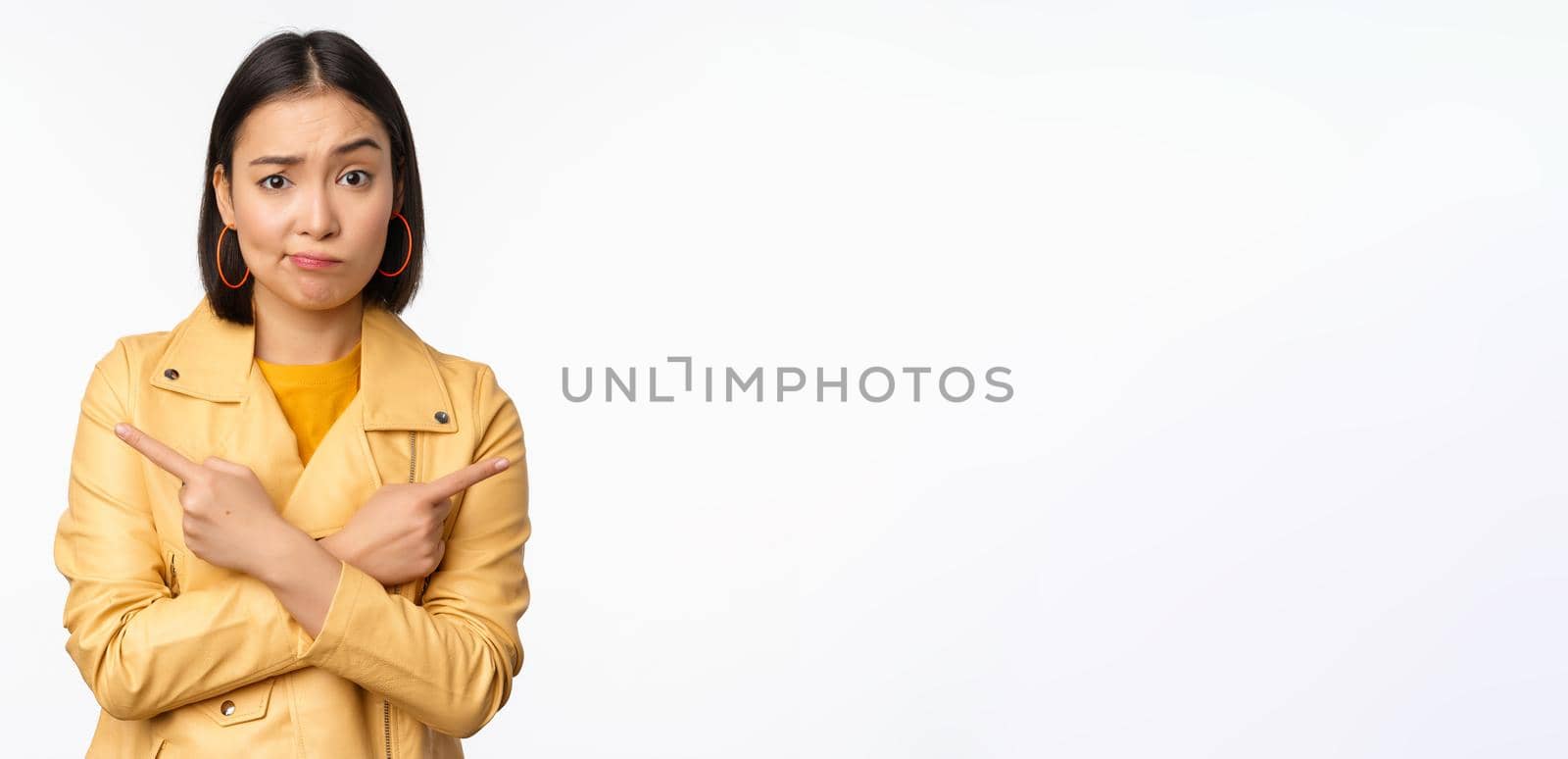 Image of indecisive asian girl, pointing fingers sideways, pointing left and right, choosing variant, deciding, standing over white background. Copy space