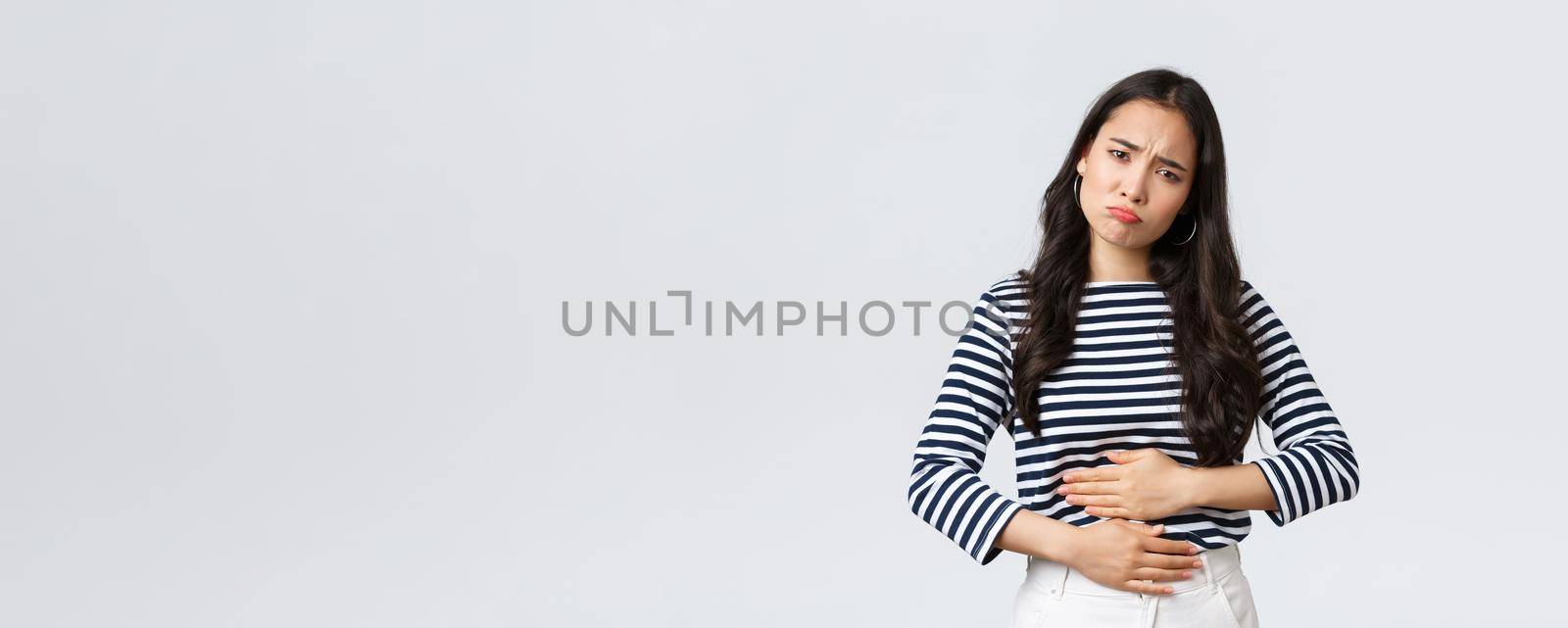 Lifestyle, beauty and fashion, people emotions concept. Woman got food poisoned, touching belly feeling unwell. Asian girl with cramps looking gloomy, having menstrual pain, white background.