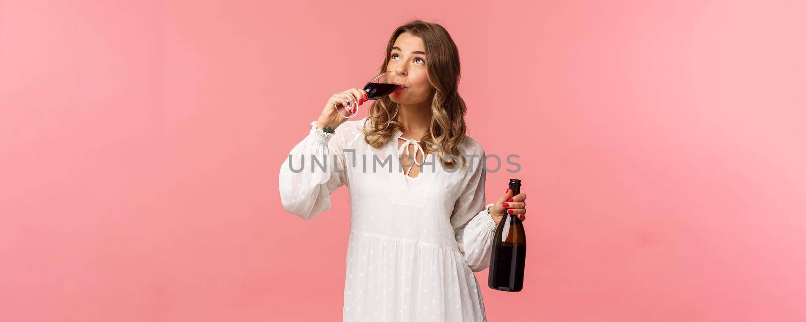 Holidays, spring and party concept. Portrait of young elegant happy blond woman, wear white trendy dress, drinking wine from glass looking up pleased, tasting good drink, hold bottle.