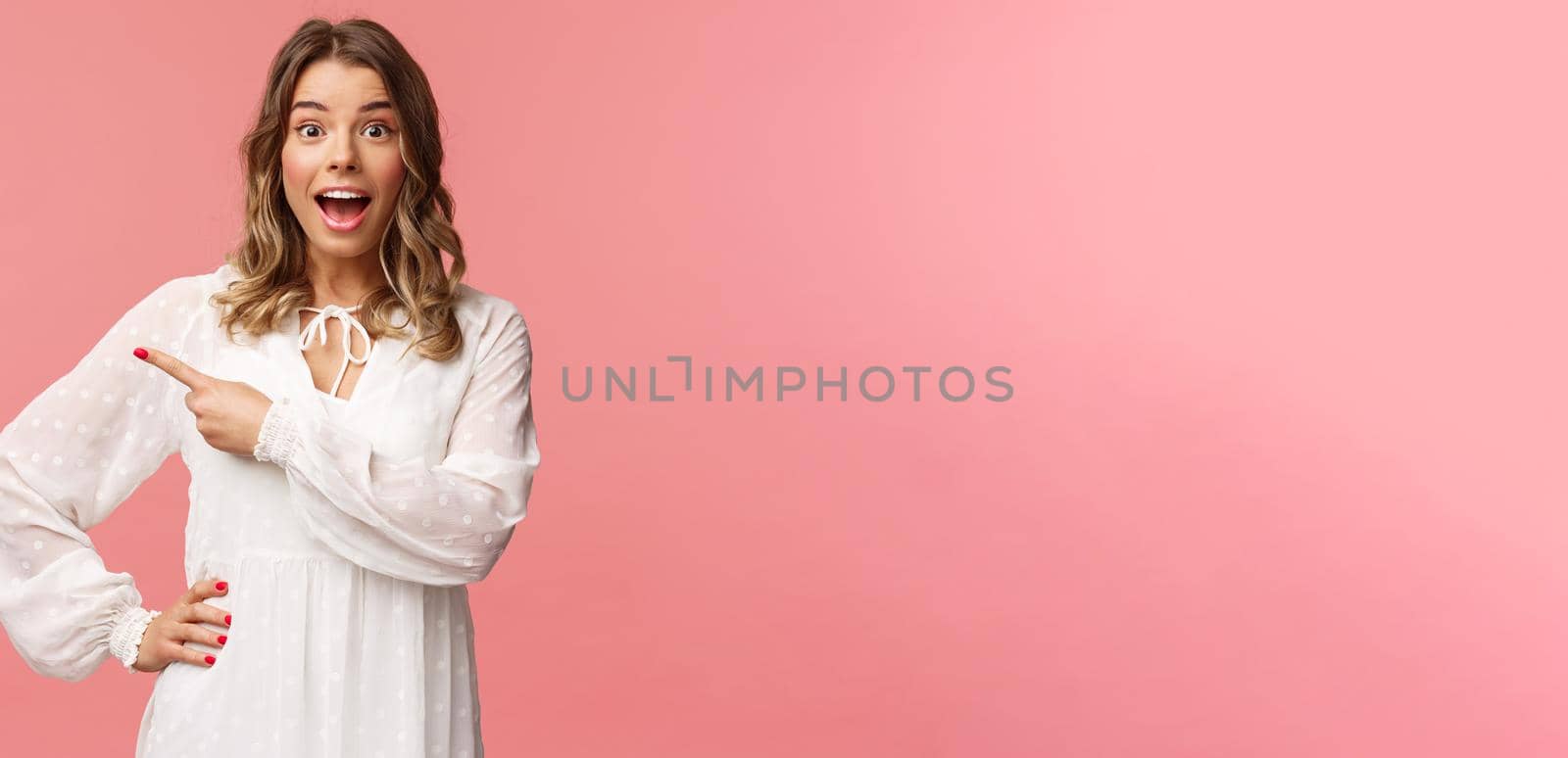 Excited and amused, intrigued blond european girl in white cute spring dress, open mouth wondered and amazed of hearing awesome news, pointing finger left at something cool, pink background.