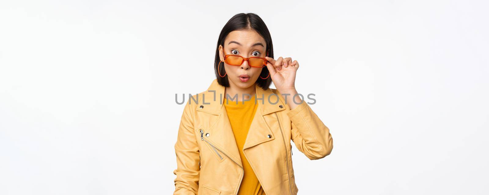 Portrait of asian brunette woman in stylish sunglasses, looks surprised and impressed at camera, checking out big news, wow face expression, standing over white background.