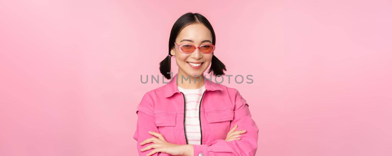 Stylish asian girl in pink clothes and sunglasses, smiling and looking happy at camera, standing over studio background.