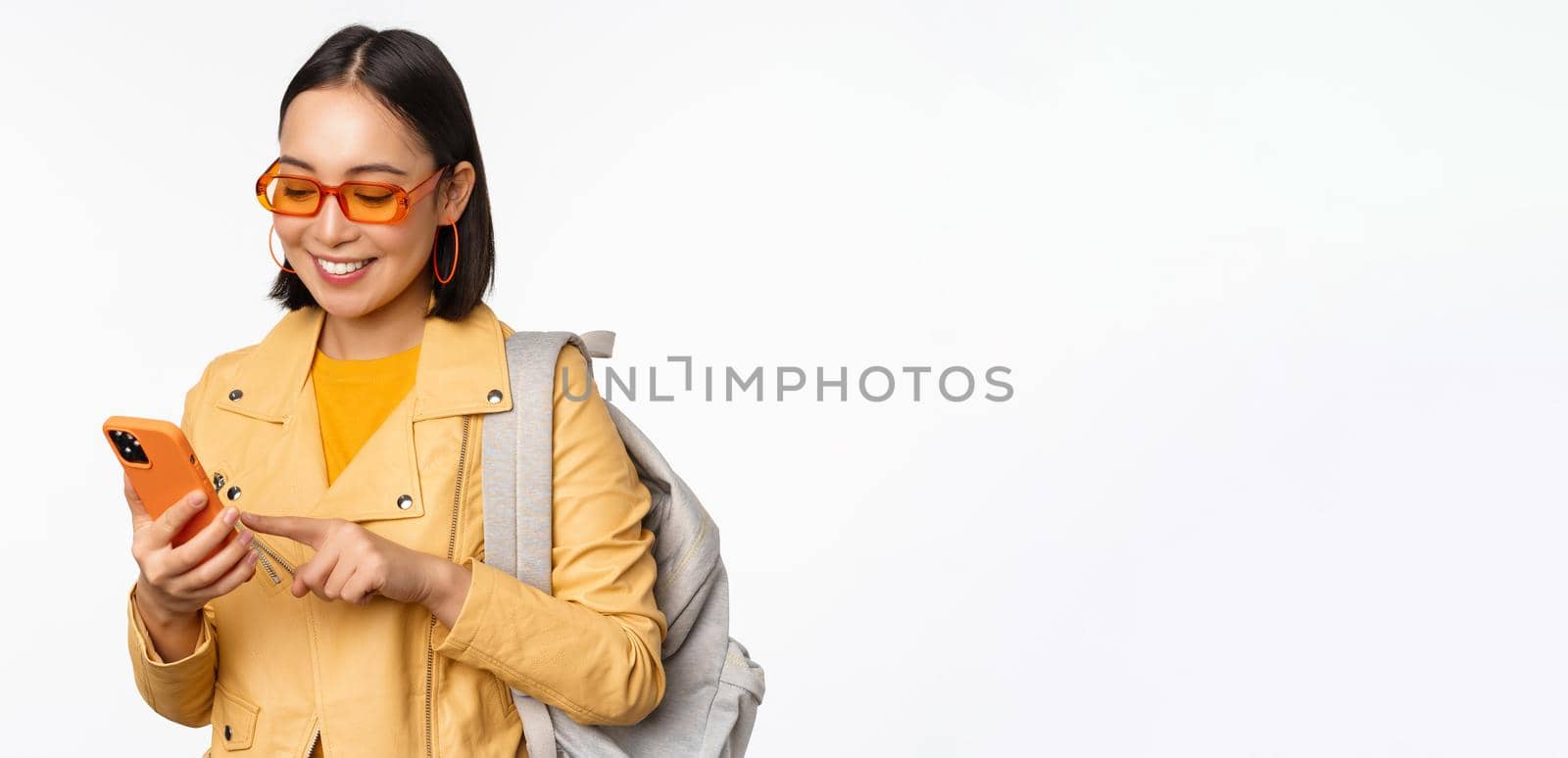 Stylish young asian woman tourist, traveller with backpack and smartphone smiling at camera, posing against white background by Benzoix